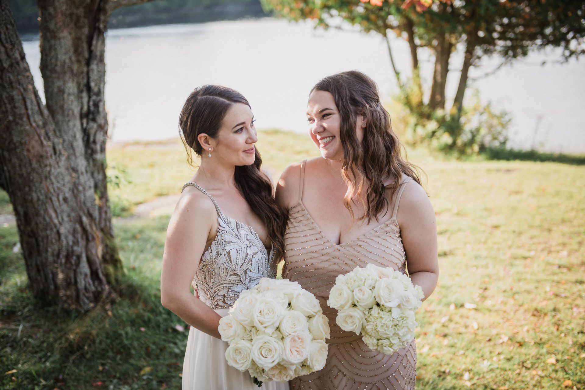 Two women in dresses holding white bouquets outdoors, smiling beside a tree in a sunny garden.