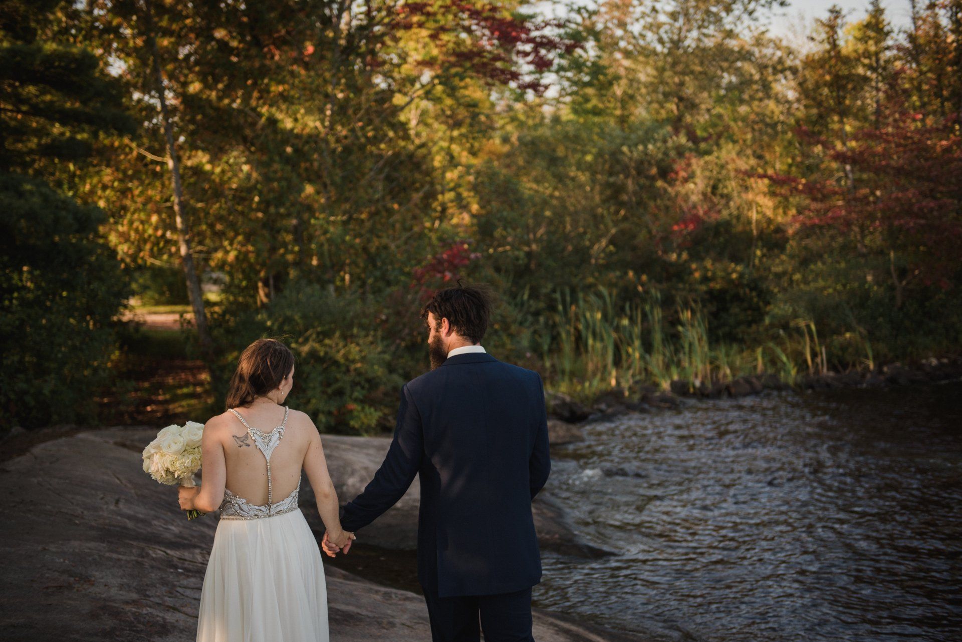 Couple holding hands by a river in autumn, with golden trees and a wedding bouquet