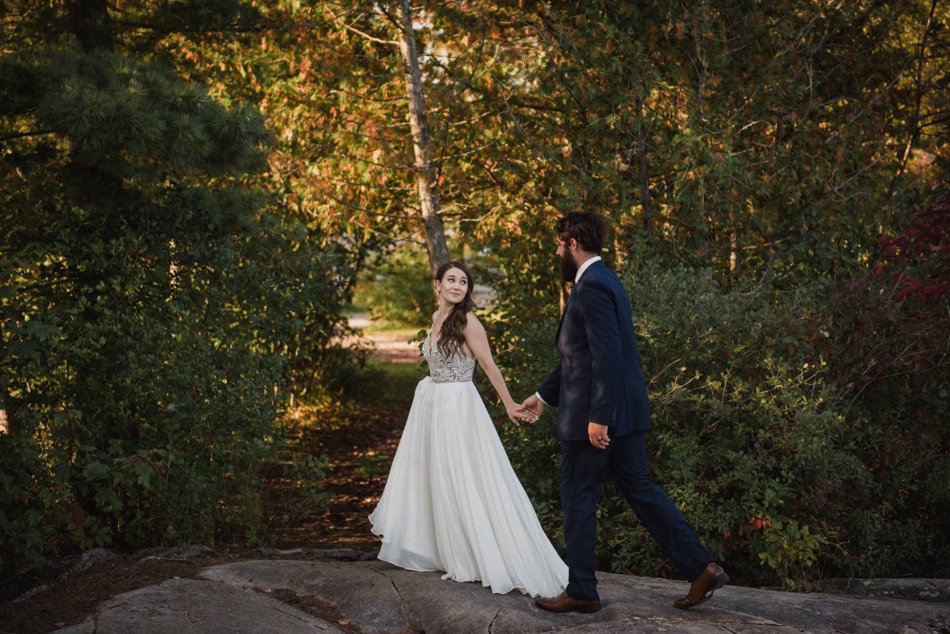 Bride and groom walking hand in hand in a wooded garden, she in a white gown and he in a dark suit.