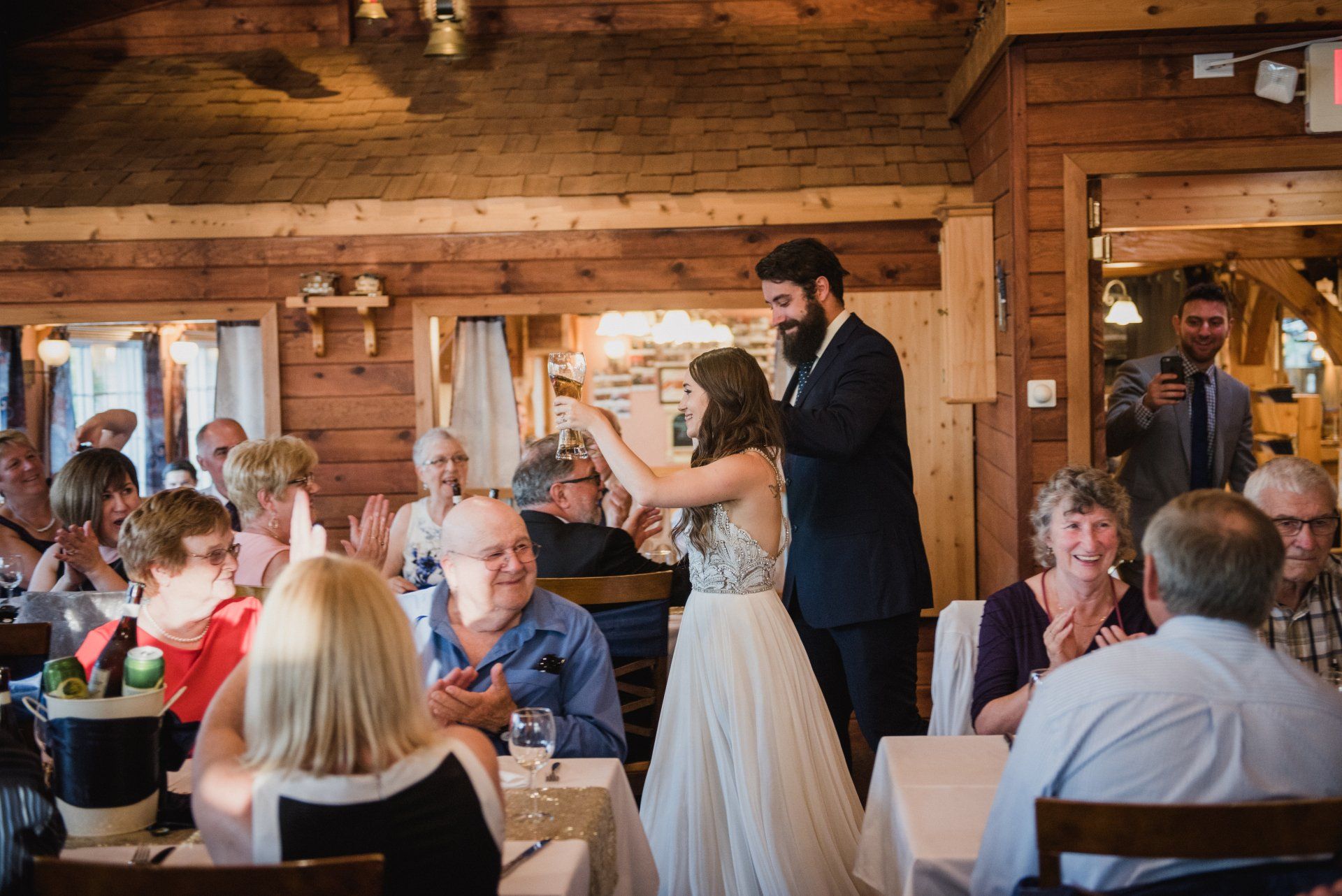 Guests gather at a rustic wood-paneled reception as a man gives a toast beside a smiling woman.