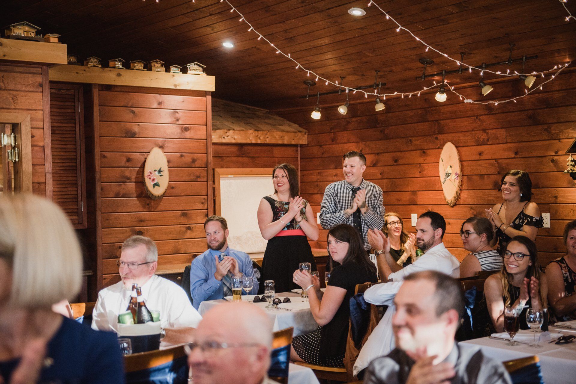 People seated and standing at a warmly lit indoor gathering in a rustic wooden hall