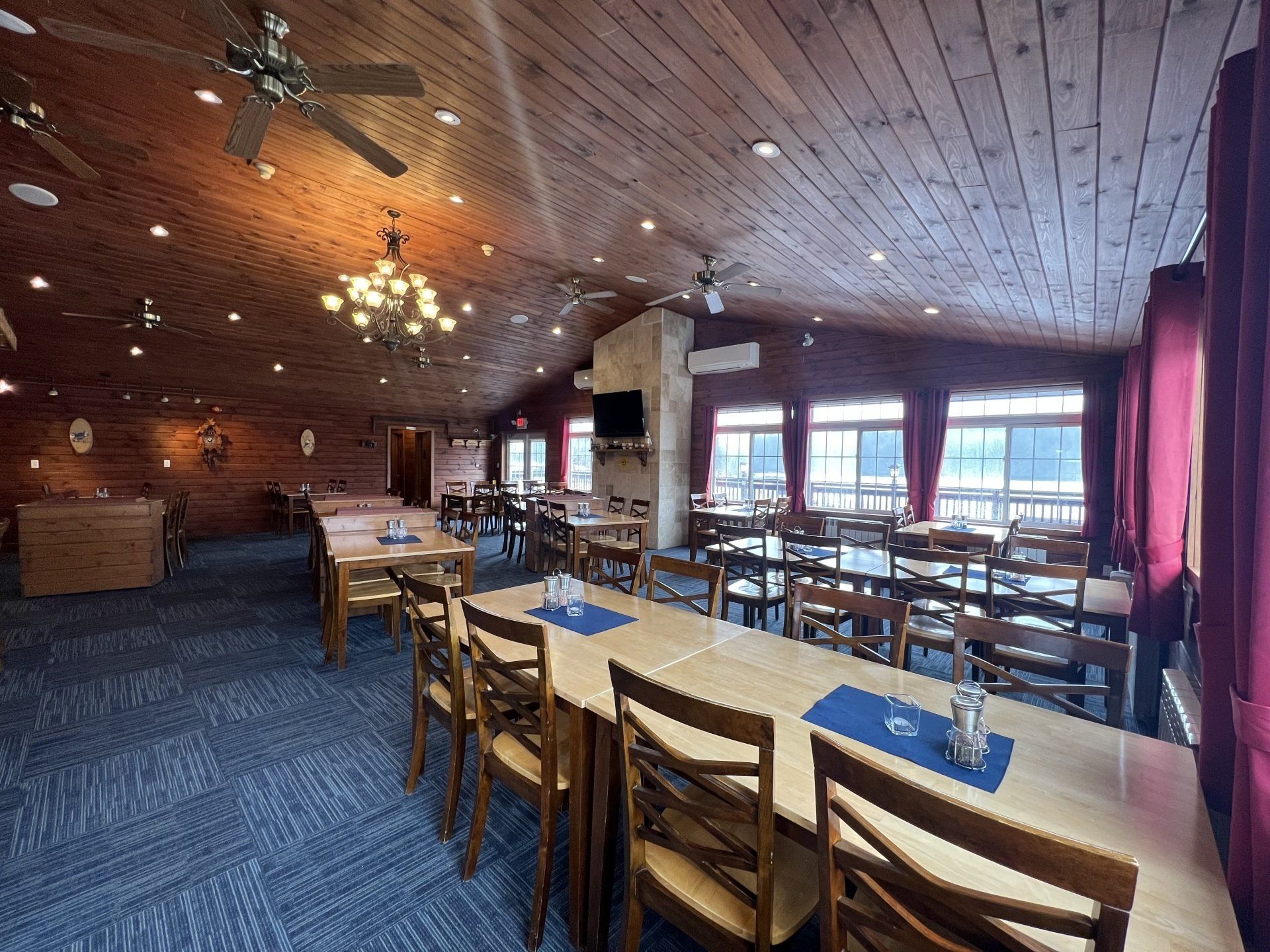 Rustic restaurant dining room with wooden tables, blue carpet, and large windows looking out over the water