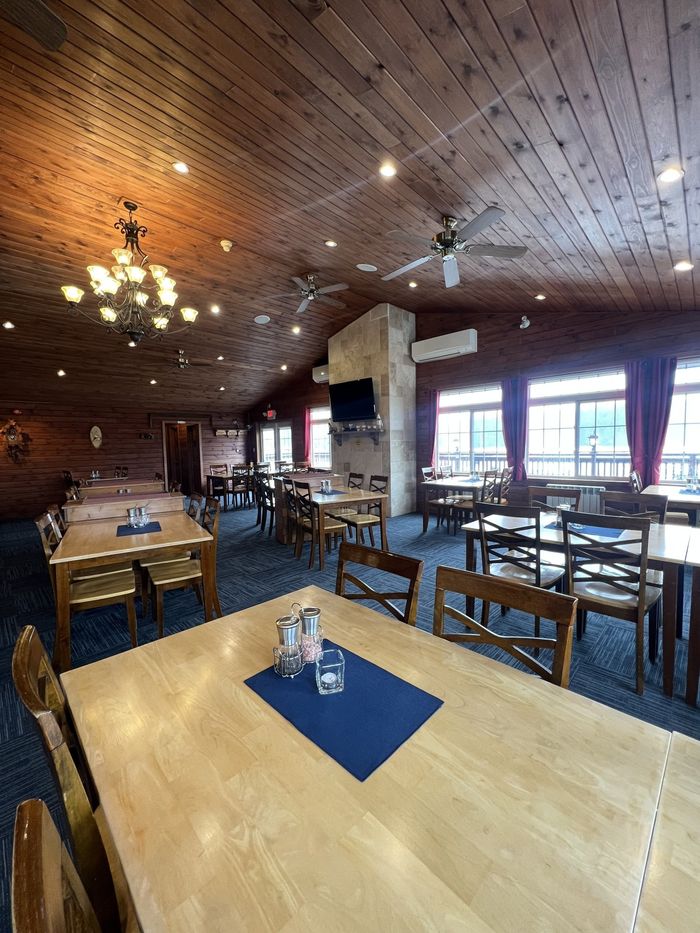 Empty restaurant dining room with wooden tables, chairs, pendant lights, and large windows.