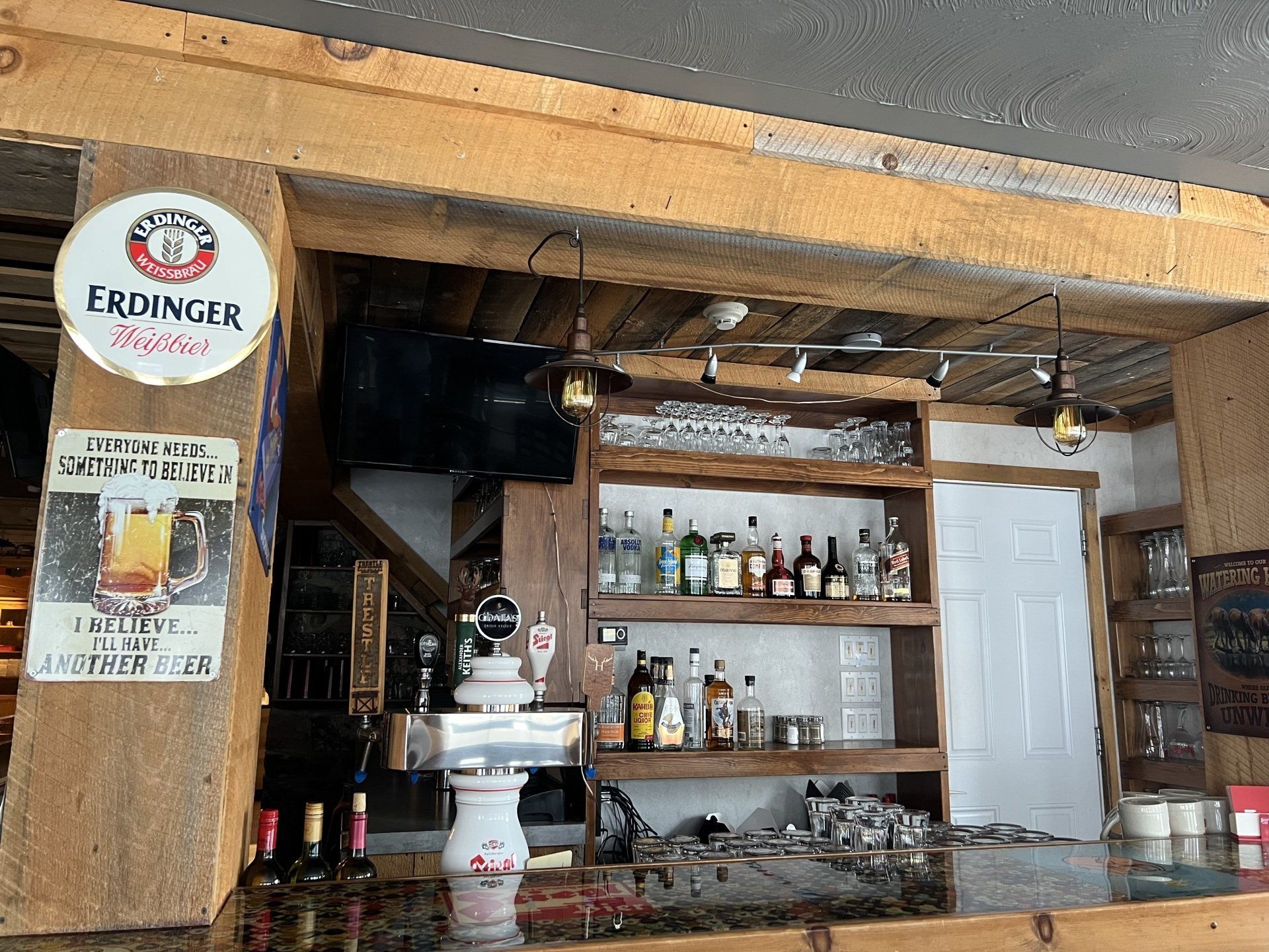 Rustic bar interior with wooden shelves, liquor bottles, white door, and a small sink counter