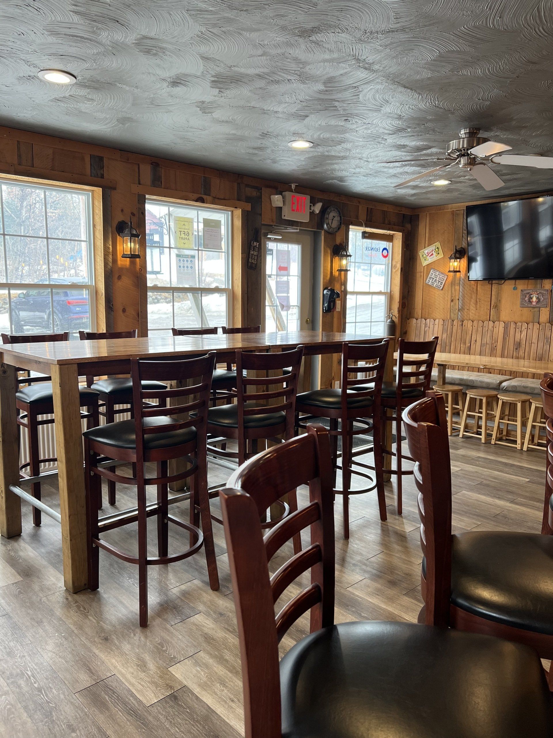 Empty bar interior with wooden tables, high stools, and large windows letting in daylight.