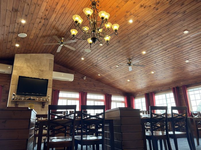Empty restaurant dining room with wooden ceiling, chandelier, tables, chairs, and a wall-mounted TV