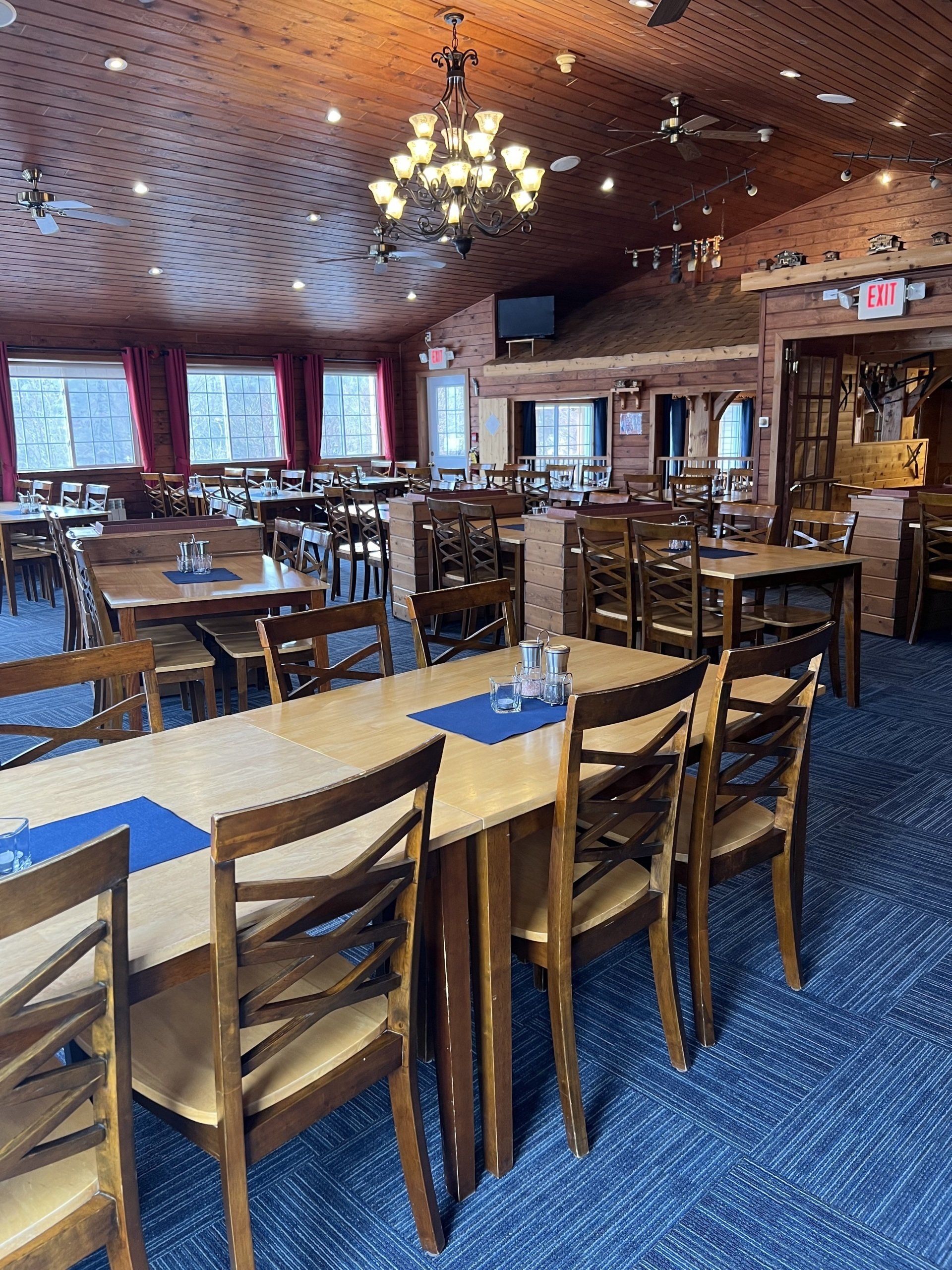 Empty restaurant dining room with wooden tables, chairs, blue carpet, and warm ceiling lights.