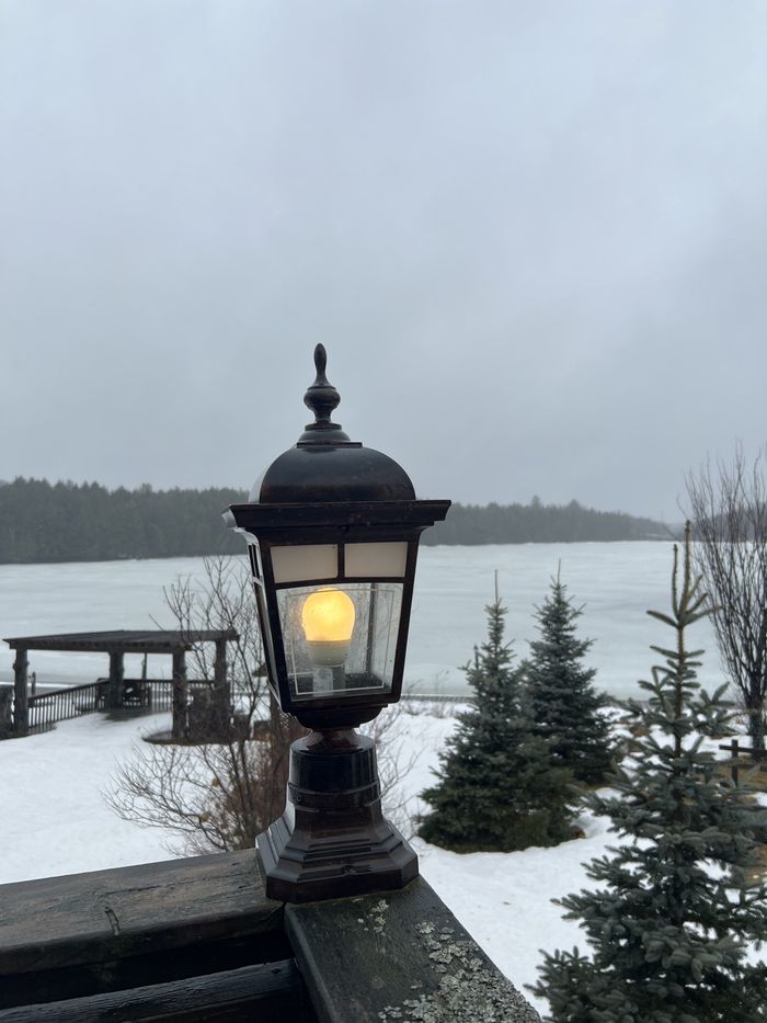 Lit black lantern glowing by a snowy lakeside with dock and evergreens under a gray sky