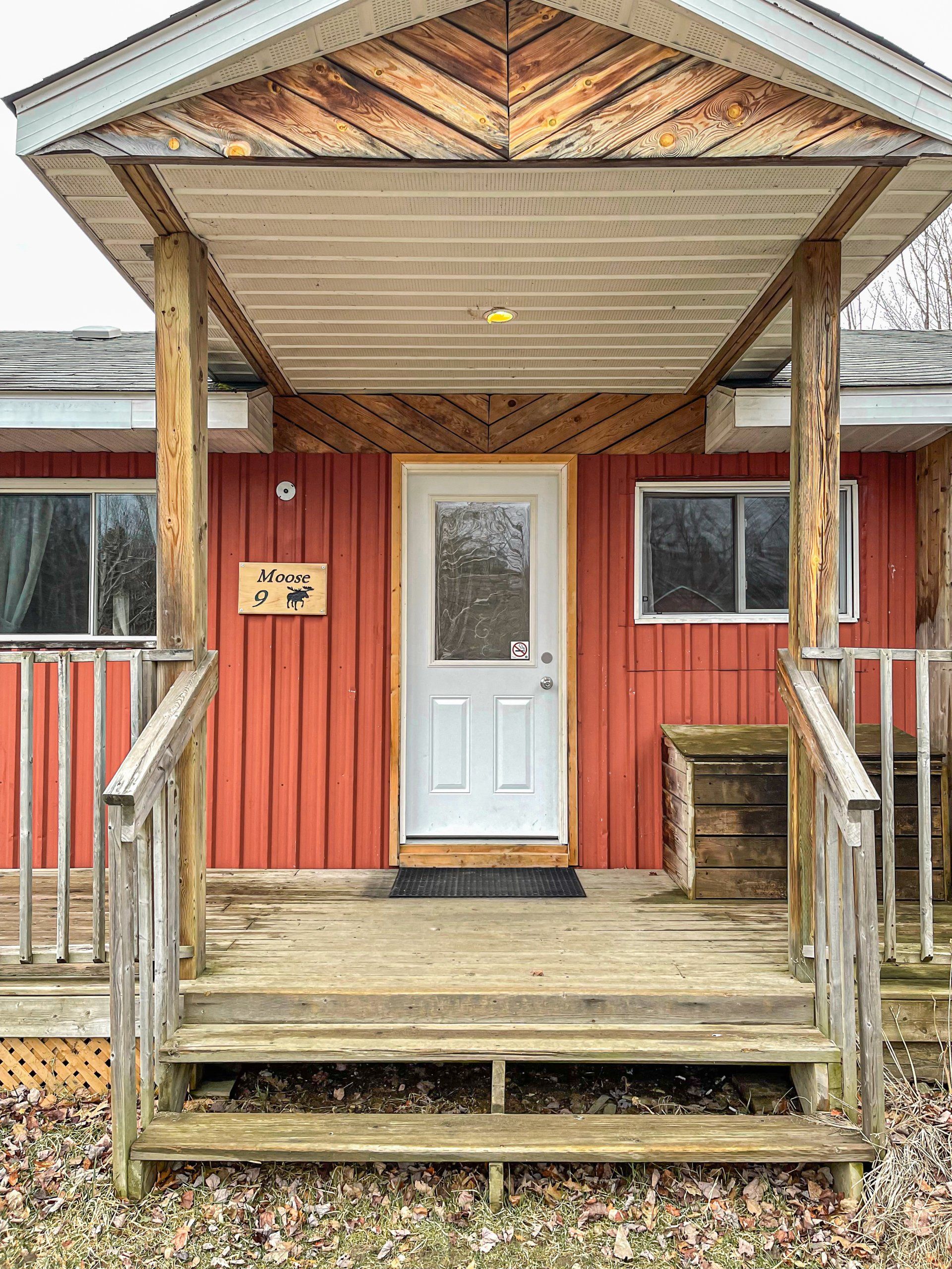 Red cabin porch with white door, wooden steps, and porch railing under a covered roof