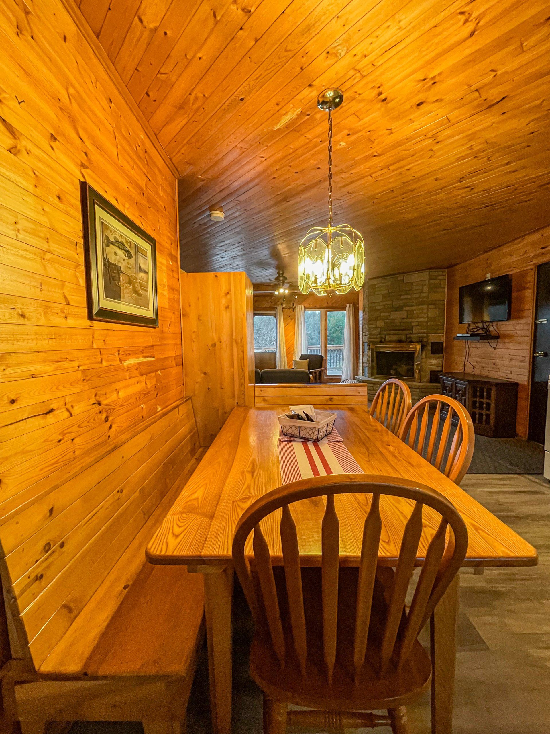 Wood-paneled dining room with a long wooden table, chairs, chandelier, and stone fireplace.