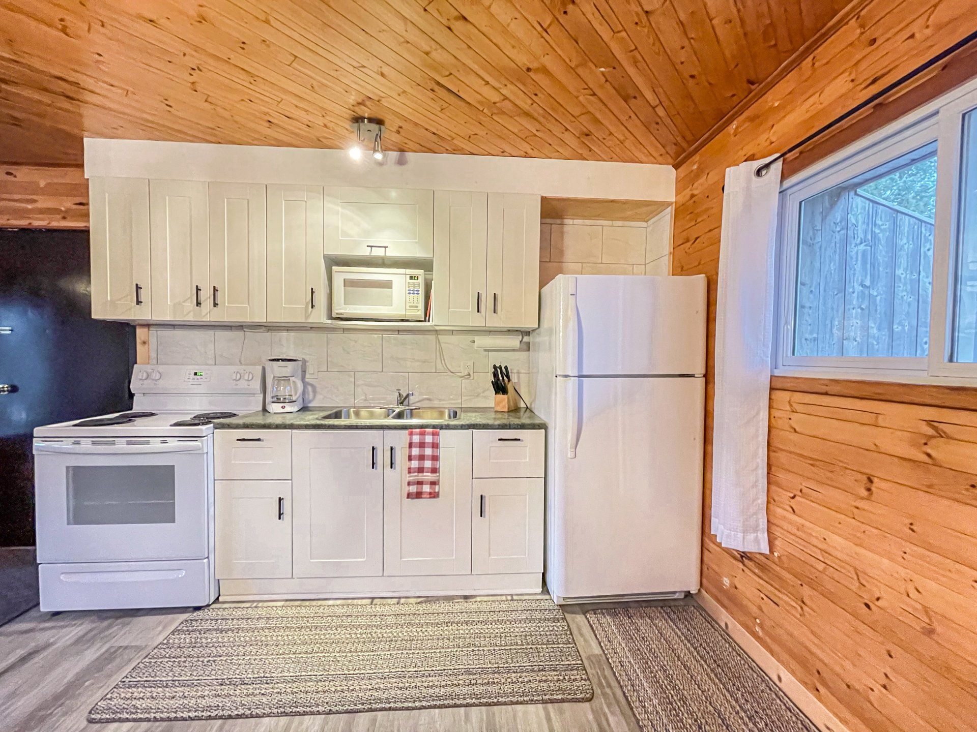 Small kitchen with white appliances, cabinets, wood-paneled walls and ceiling, and a patterned rug.