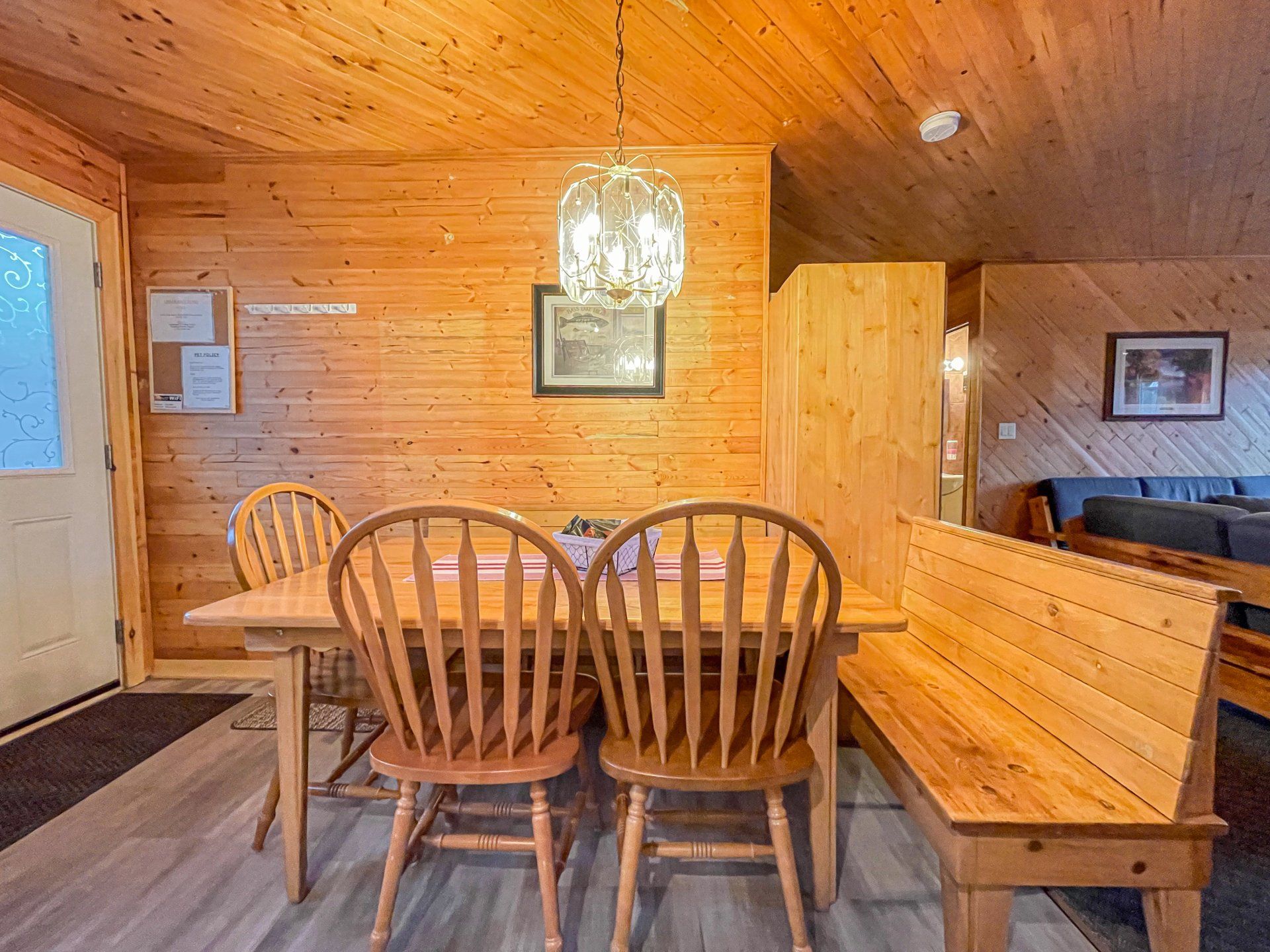 Wood-paneled dining room with a wooden table, chairs, bench, chandelier, and framed wall art.