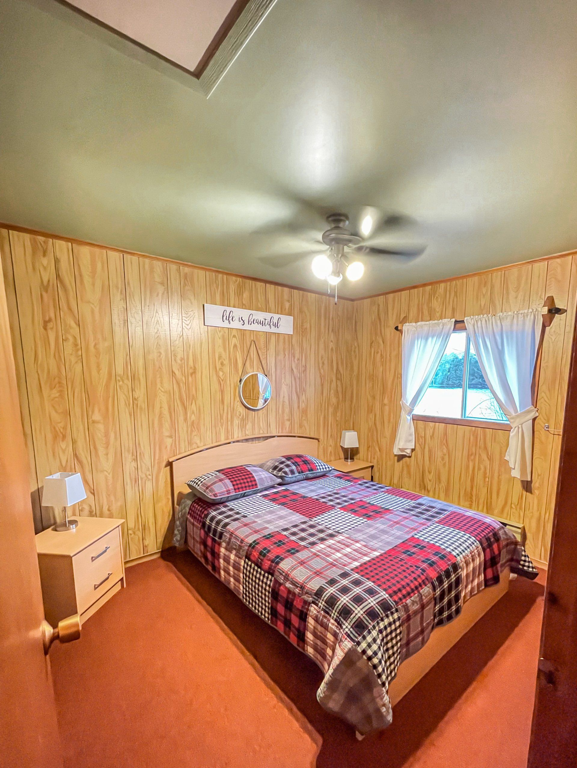 Small bedroom with wood-paneled walls, red carpet, plaid bedspread, ceiling fan, and window with curtains