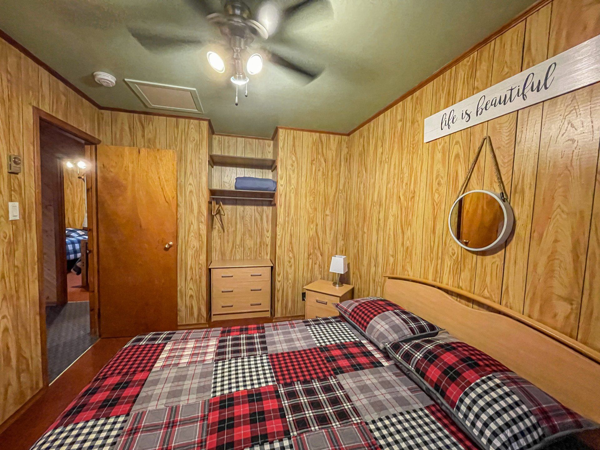 Cozy wood-paneled bedroom with plaid bedspread, mirror, closet, and ceiling fan