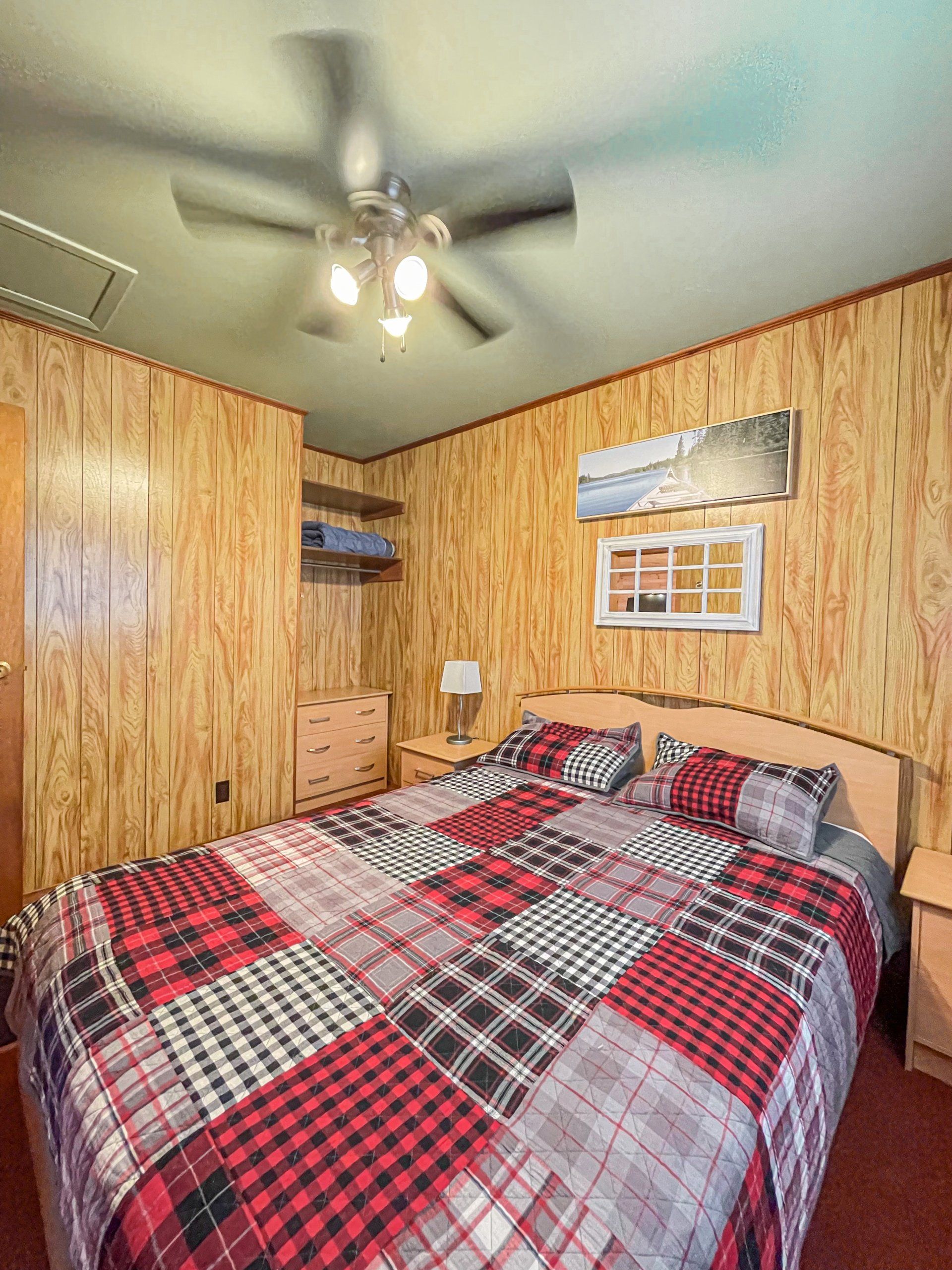 Cozy bedroom with wood-paneled walls, plaid bedspread, ceiling fan, and small window.