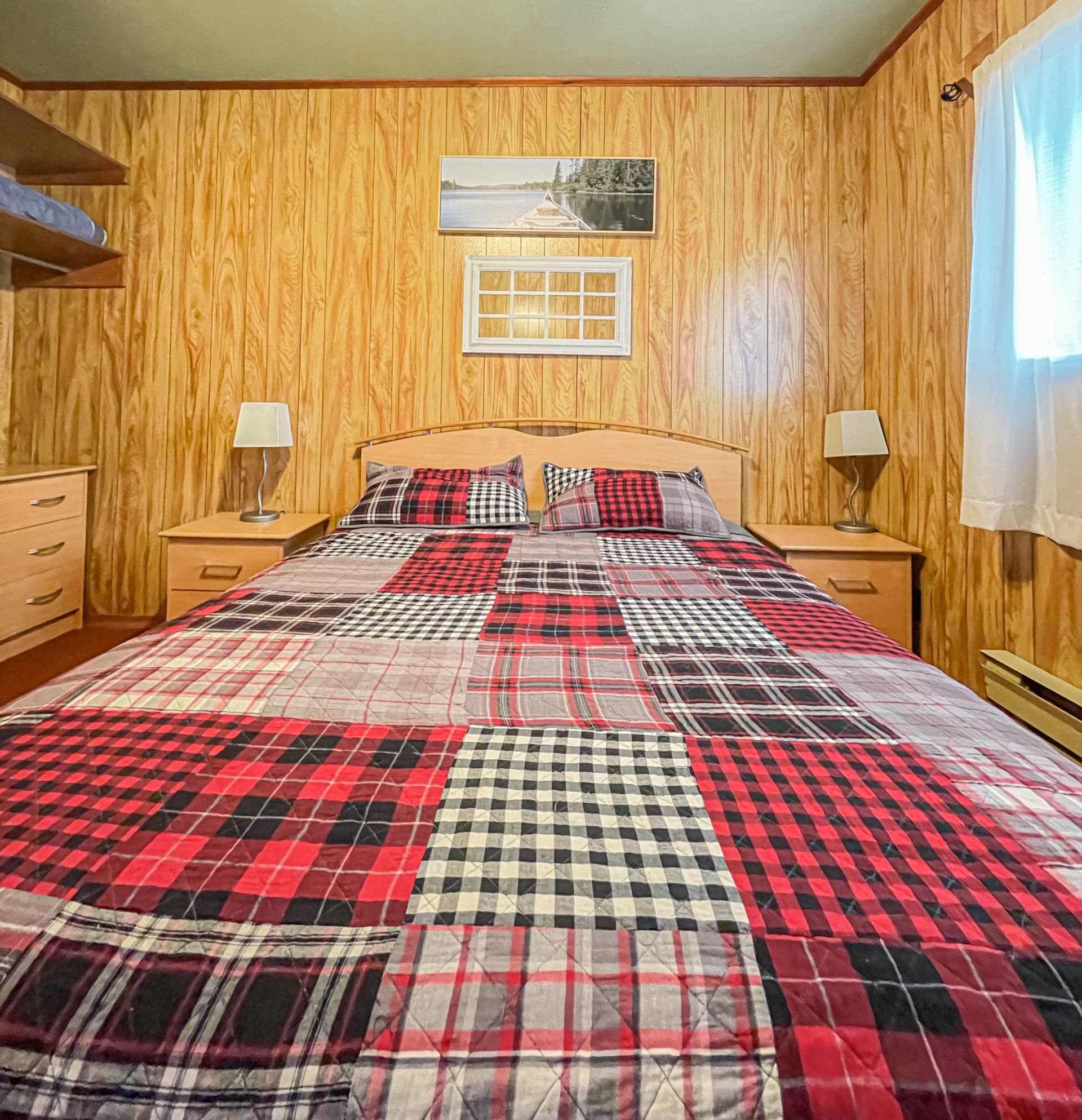 Bedroom with red plaid bedspread, wood-paneled walls, bedside lamps, and a window with blue curtains