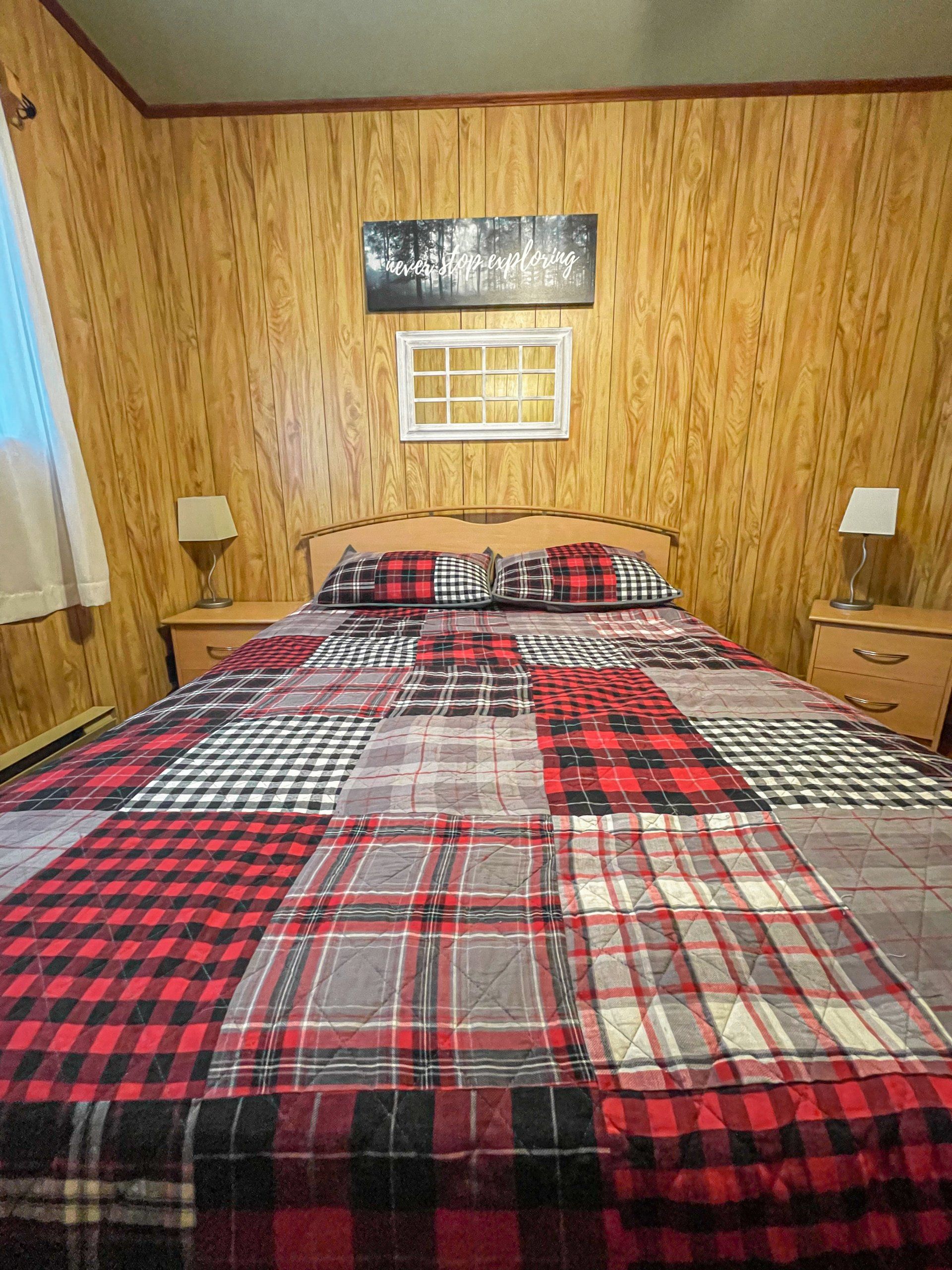 Neatly made bed with red and gray plaid blanket in a wood-paneled bedroom, with matching lamps and wall art.