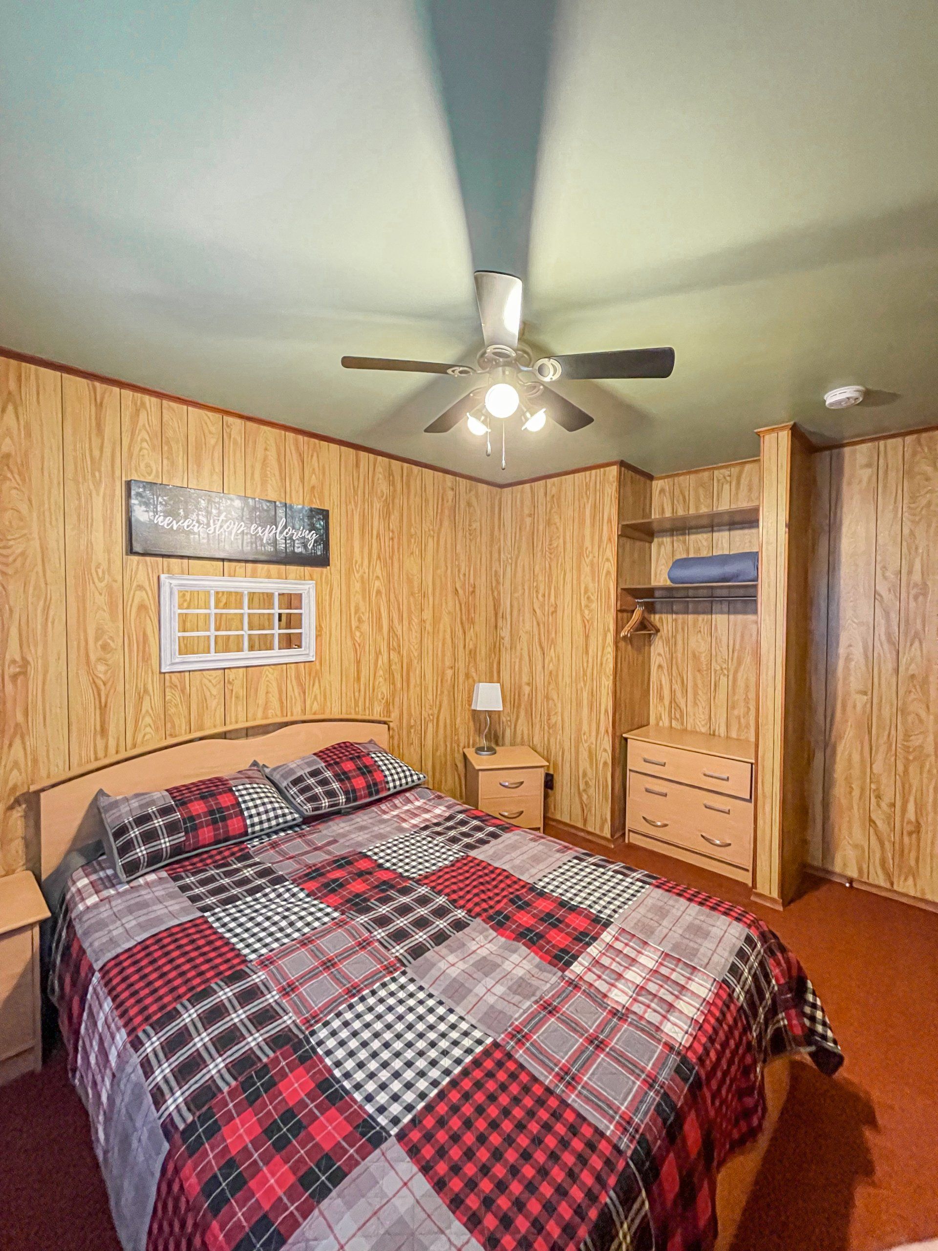 Small bedroom with wood-paneled walls, plaid bedspread, ceiling fan, and dresser near a closet.