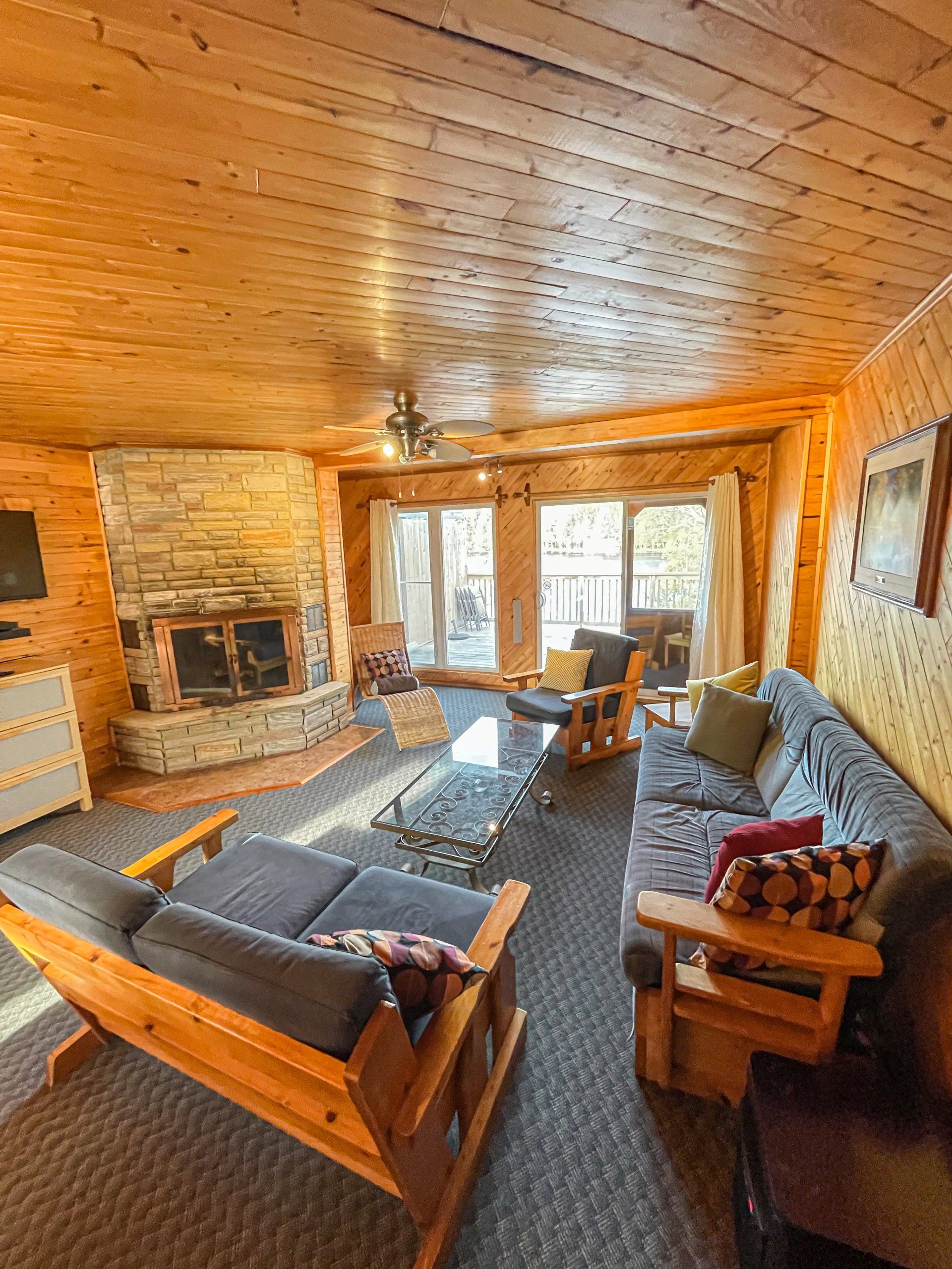 Cozy log cabin living room with stone fireplace, wooden ceiling, and large windows overlooking snowy trees