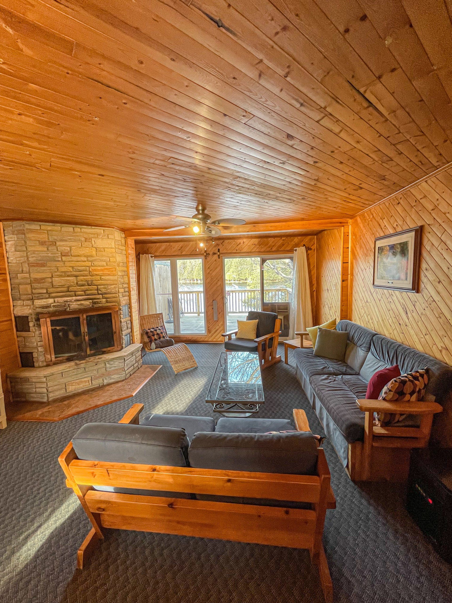 Cozy wood-paneled living room with stone fireplace, sofas, and large windows overlooking a deck