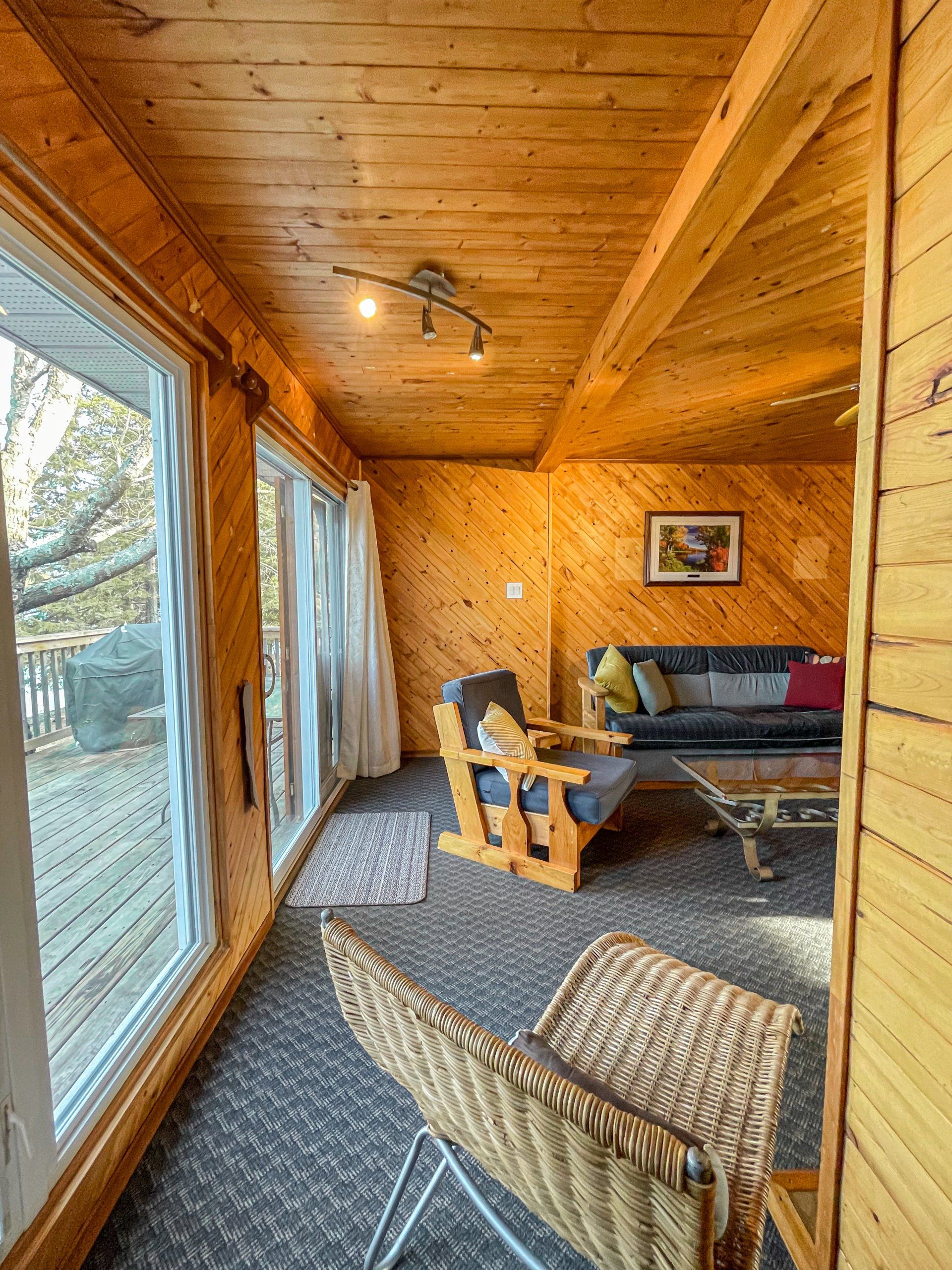 Cozy wood-paneled sunroom with large windows, wicker chair, sofa, and patterned rug