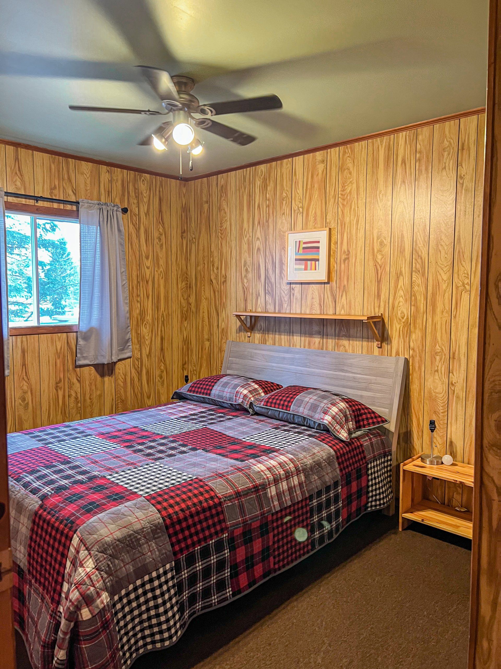 Small bedroom with a bed, ceiling fan, window, and wood-paneled walls.