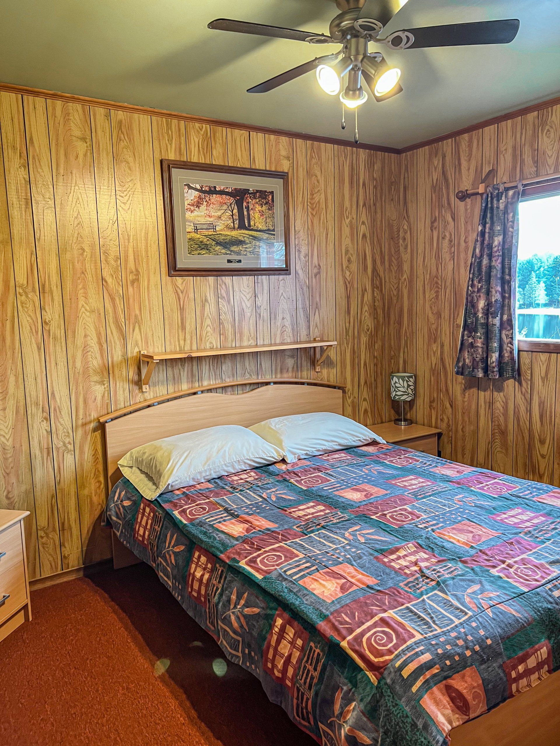 Cozy bedroom with a bed, wood-paneled walls, ceiling fan, framed art, and a window with curtains.