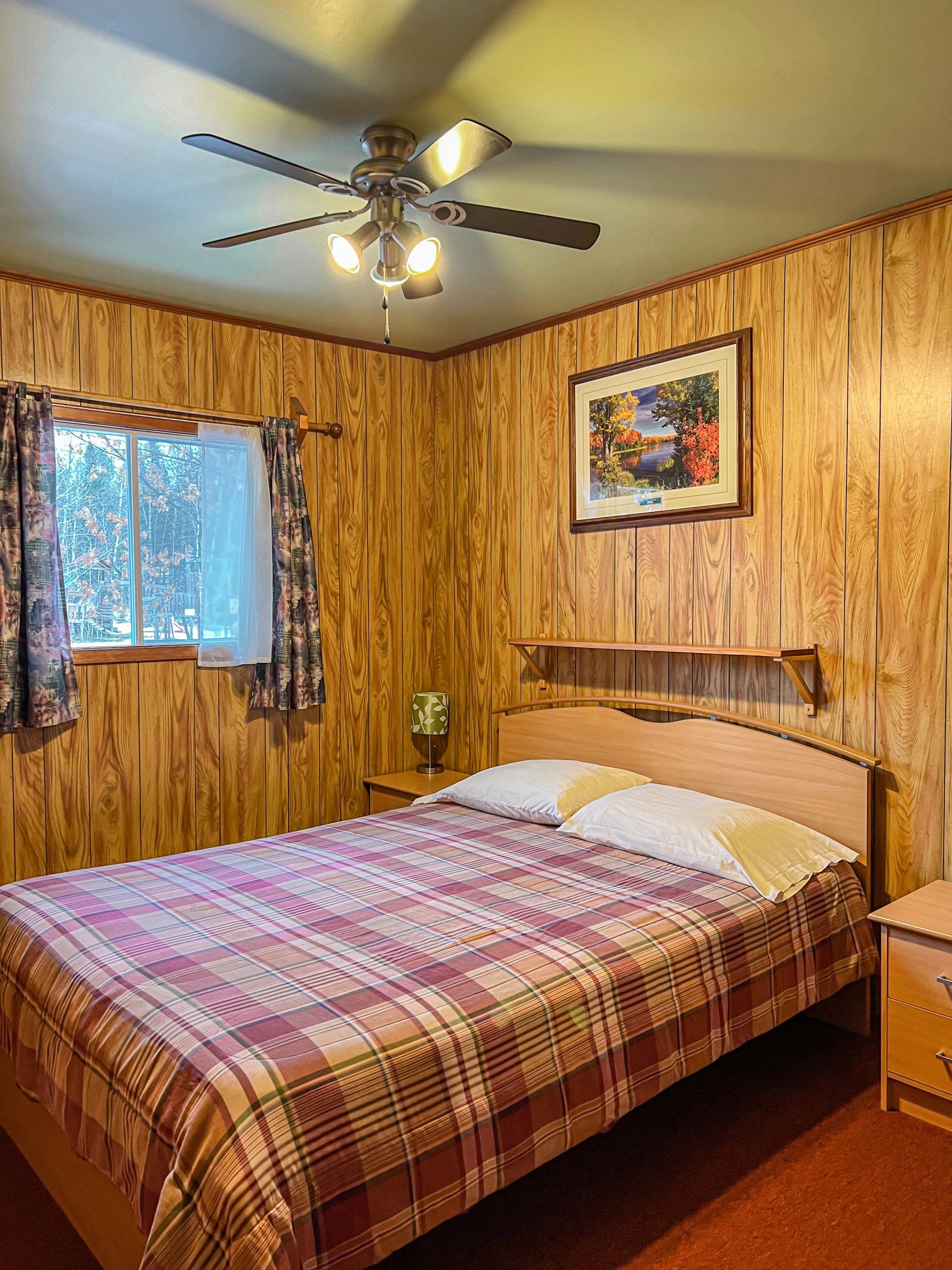 Wood-paneled bedroom with a bed, ceiling fan, window, and framed wall art