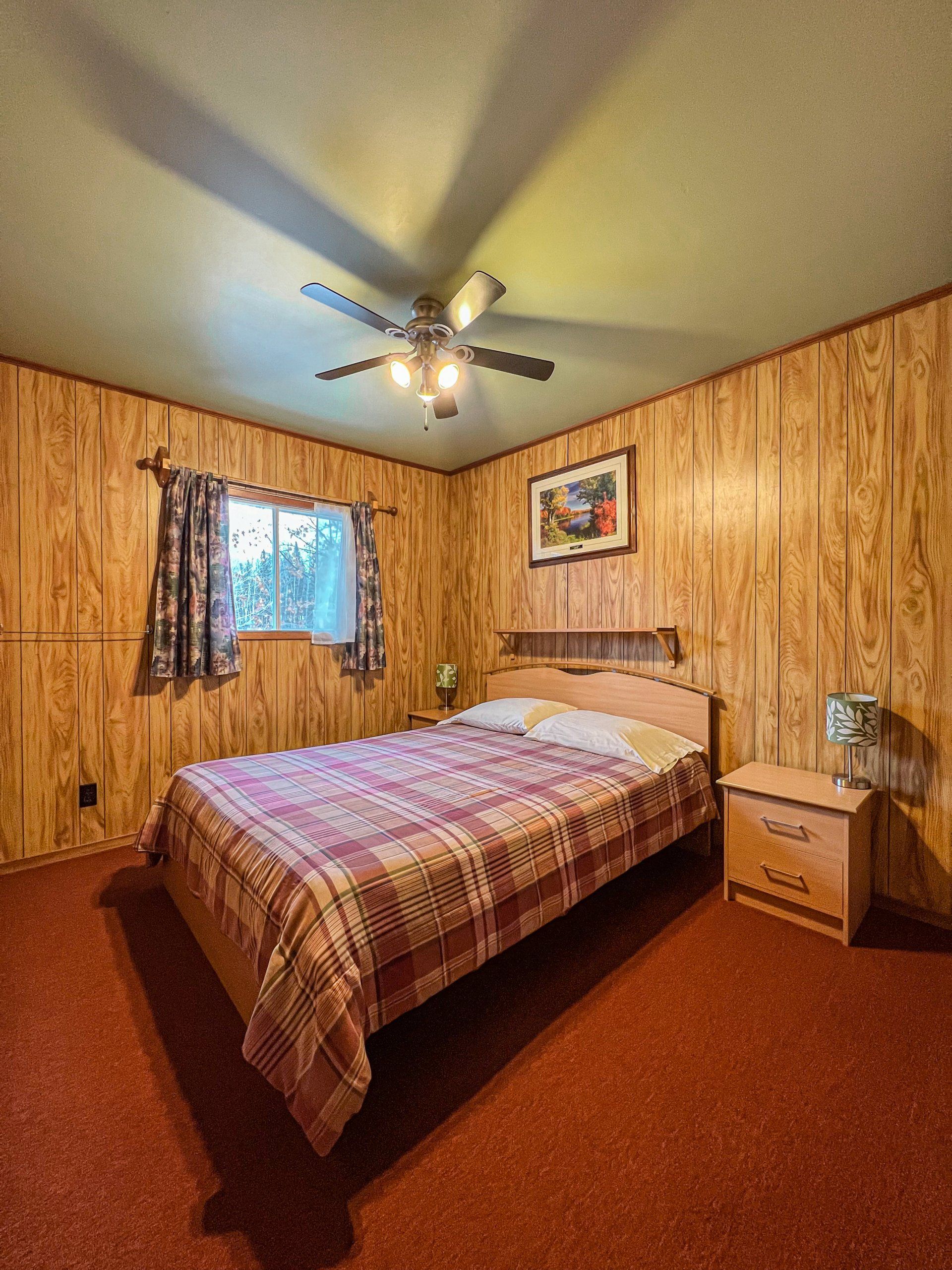 Small wood-paneled bedroom with a bed, nightstand, window, and ceiling fan-light.
