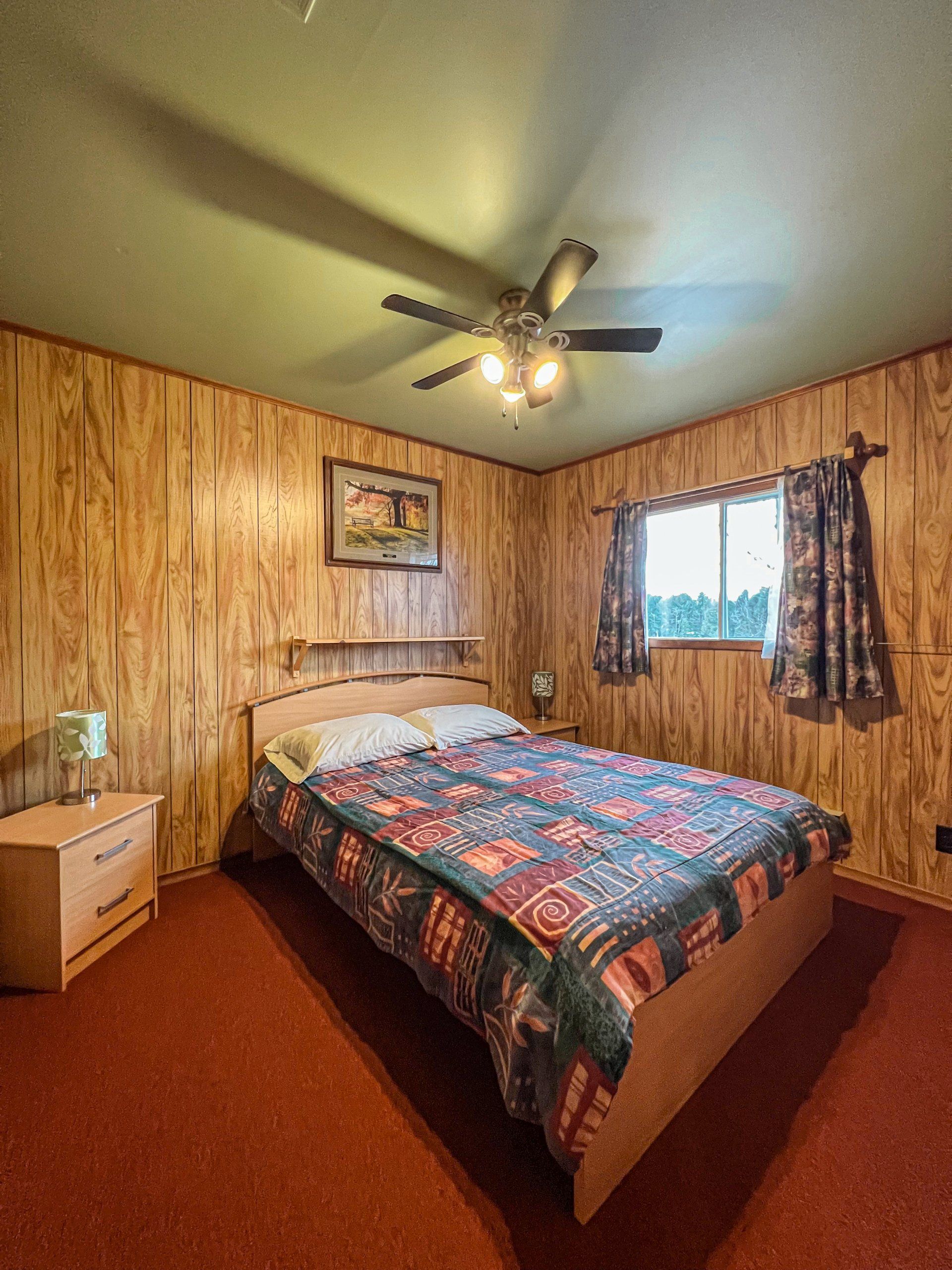 Cozy bedroom with wood-paneled walls, a bed with colorful quilt, window, nightstand, and ceiling fan.