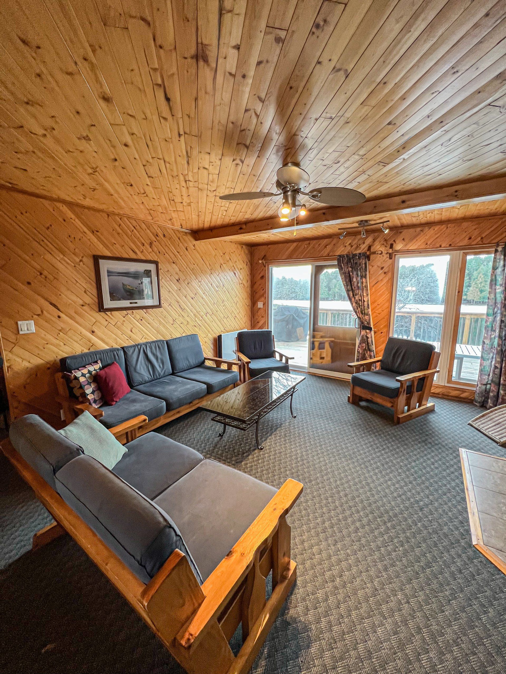 Cozy wood-paneled living room with gray sofas, armchairs, and large windows overlooking trees