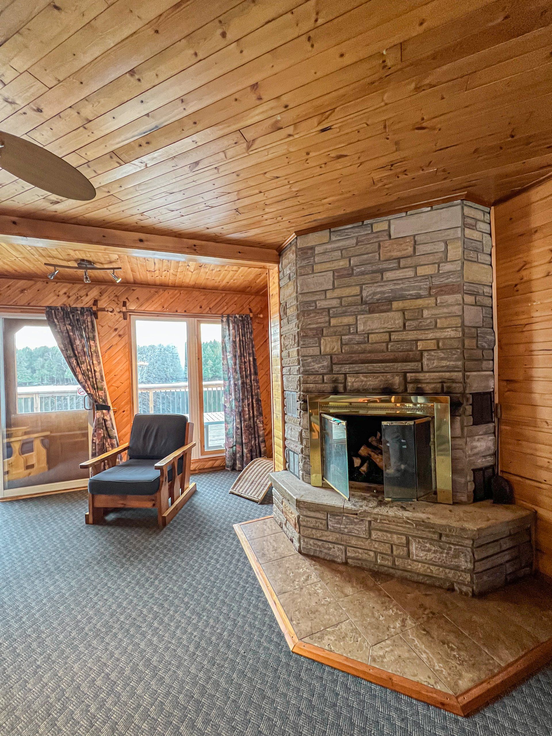 Rustic living room with stone fireplace, wood-paneled ceiling, and large windows overlooking a lake.