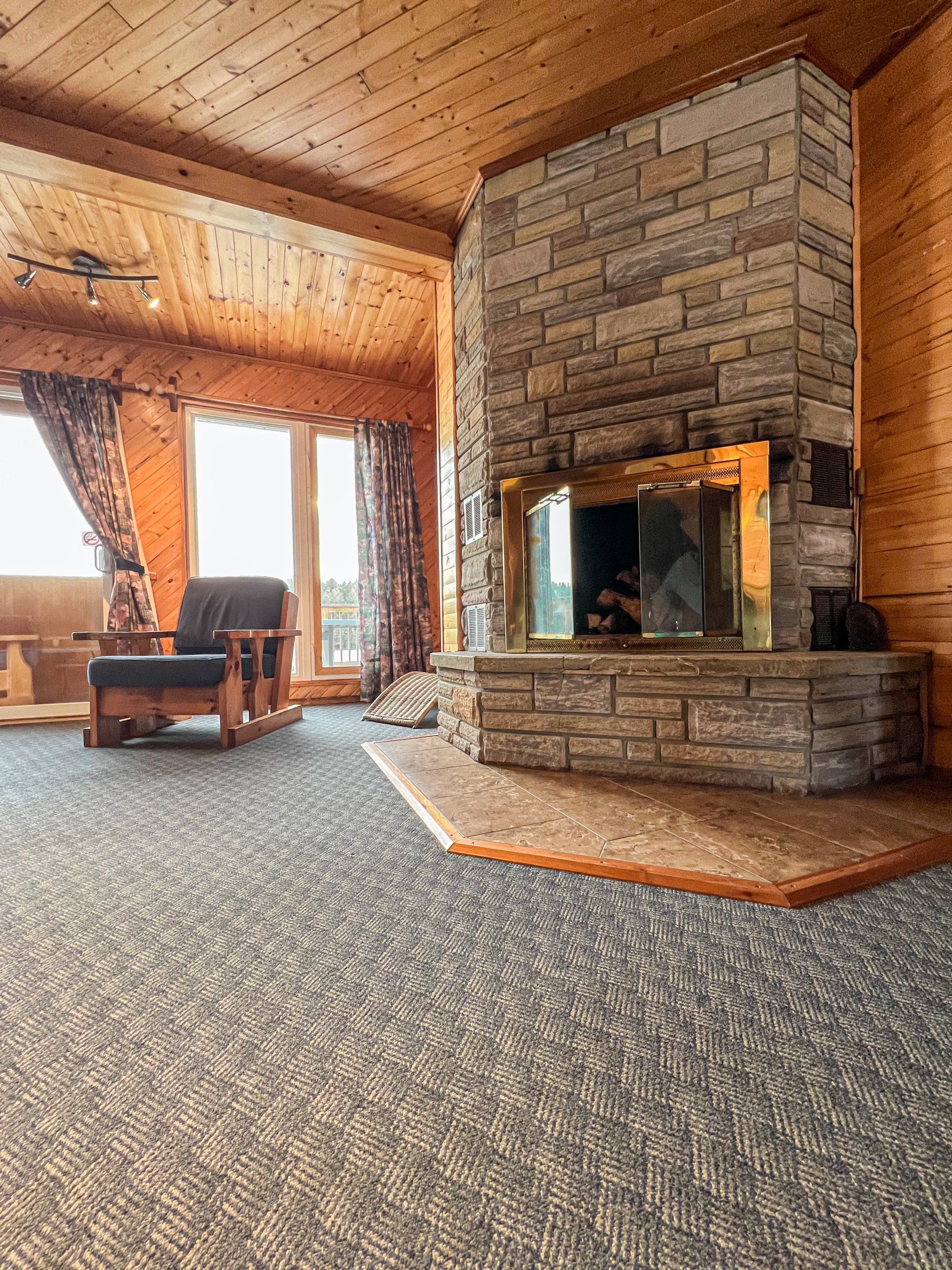 Rustic living room with stone fireplace, wood-paneled ceiling, large windows, and patterned carpet.