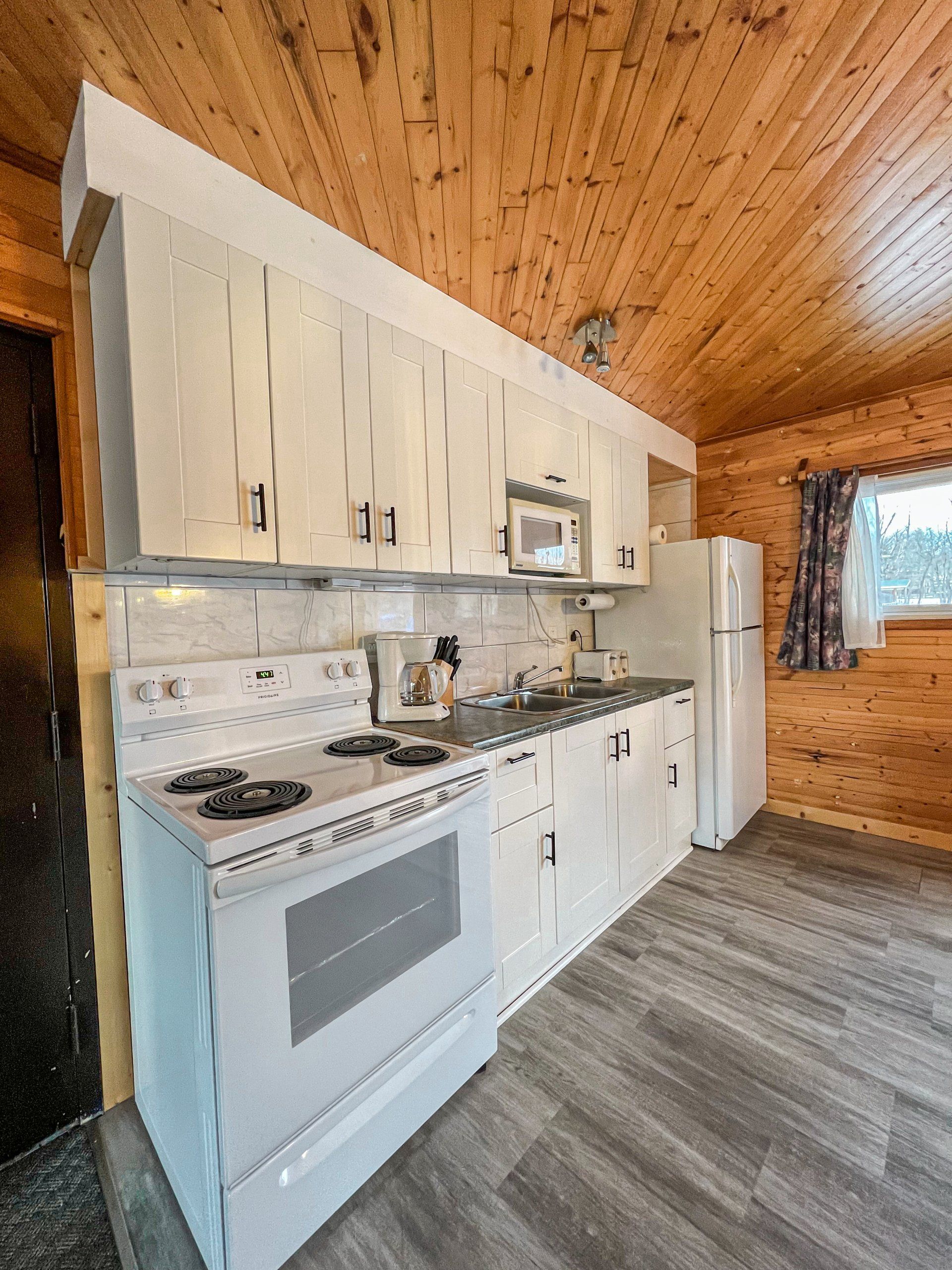 Compact white kitchen with stove, cabinets, refrigerator, and wood-paneled walls and ceiling