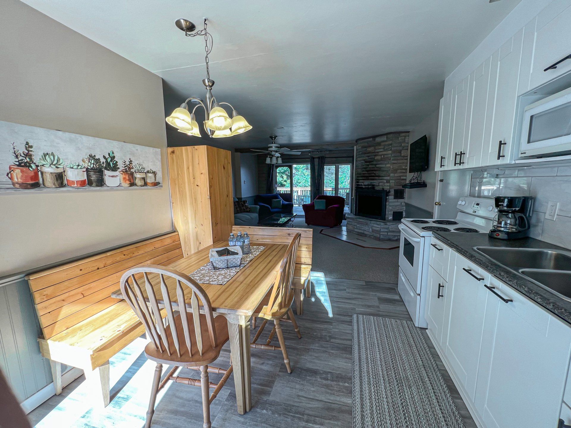 Bright dining kitchen with wooden bench, white cabinets, hanging light, and stone fireplace at the far end