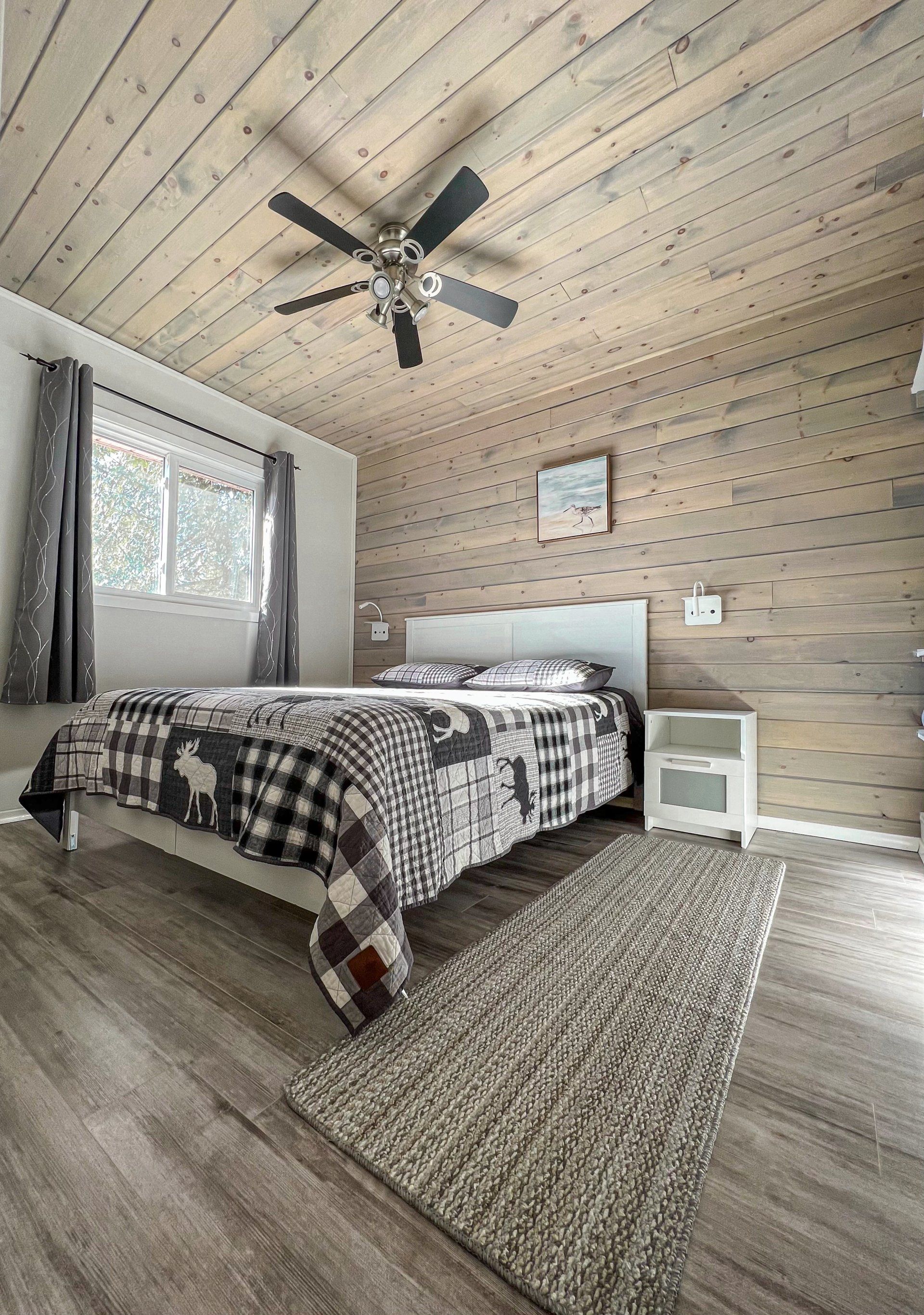 Cozy bedroom with slanted wood ceiling, bed, window, ceiling fan, and woven rug.