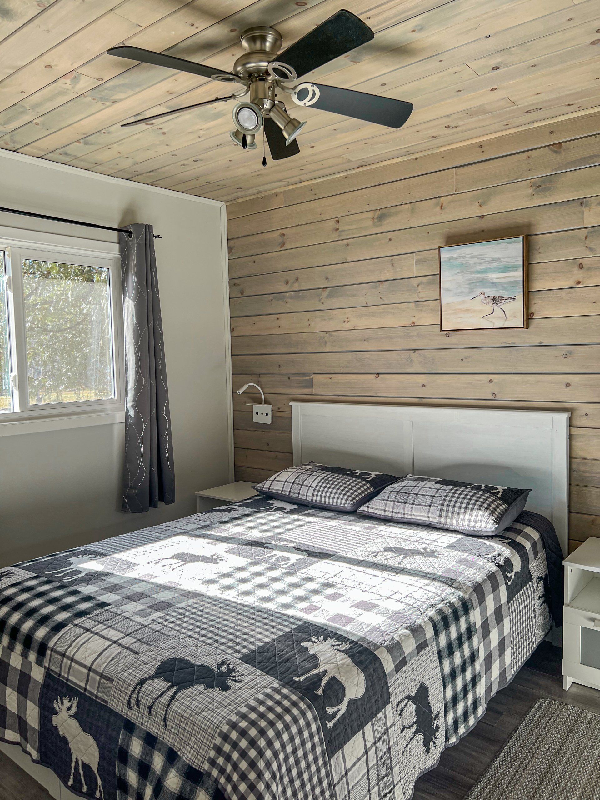 Bedroom with a made bed, ceiling fan, window, and wood-paneled wall.