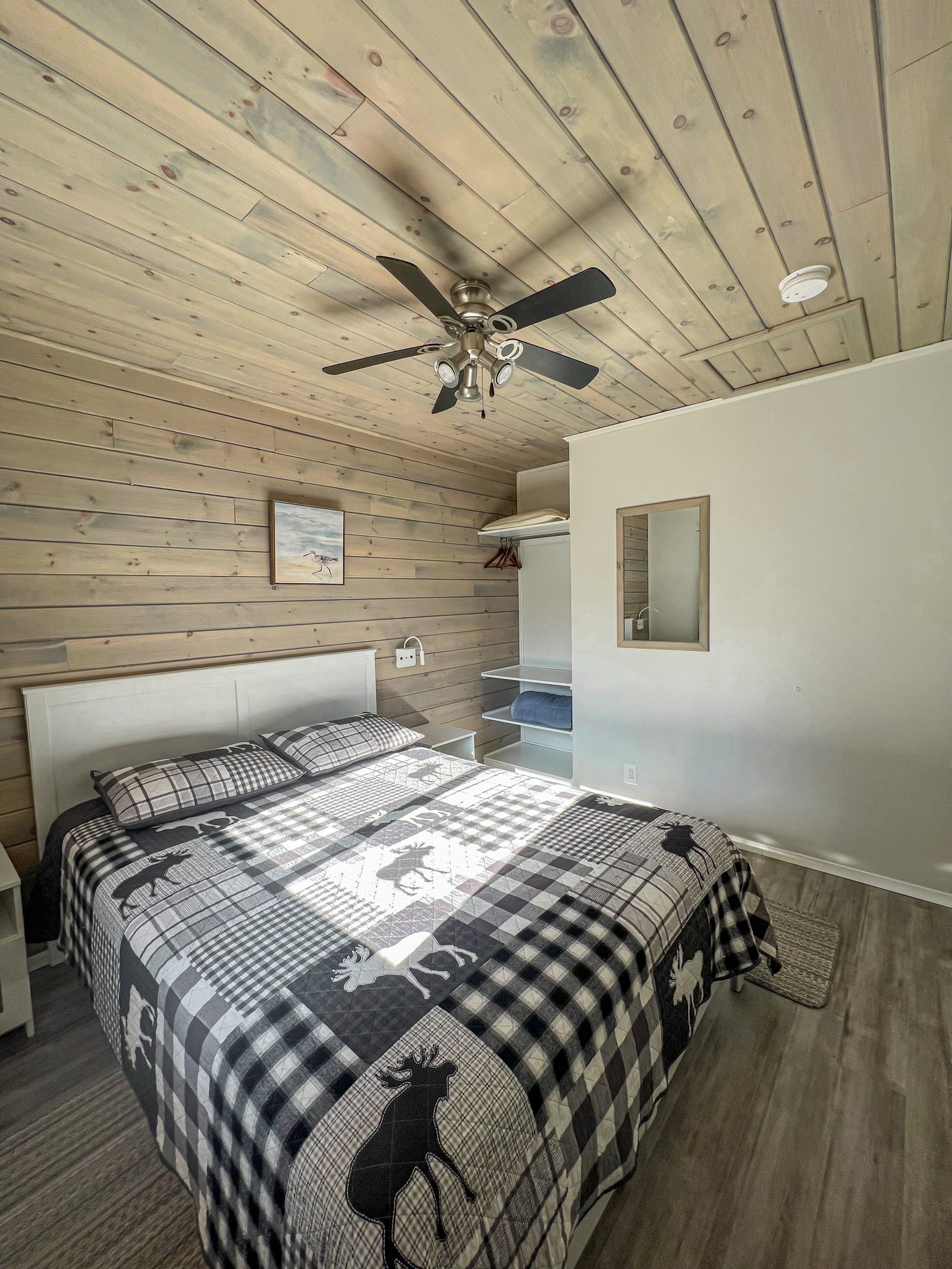 Bedroom with black-and-white quilted bed, wood-paneled walls and ceiling, and a ceiling fan above.