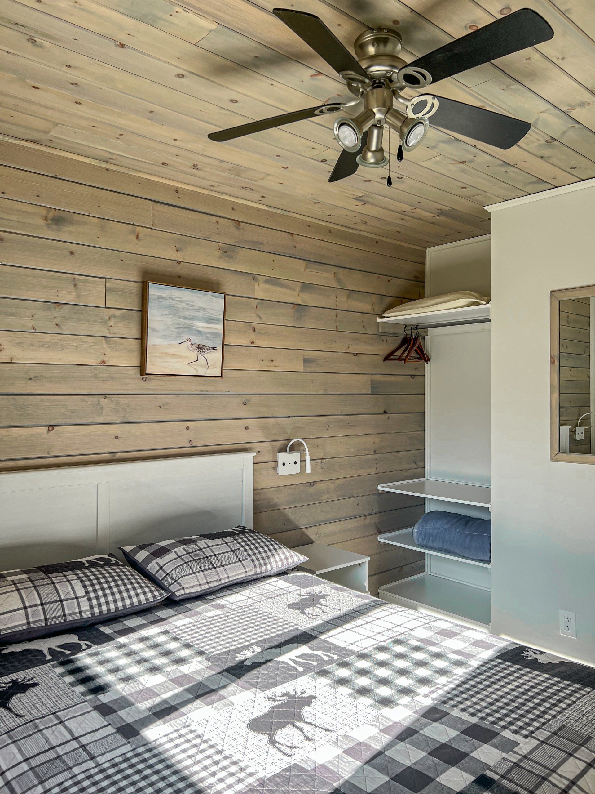Bedroom with plaid bedspread, wood-paneled walls, ceiling fan, and open closet shelves.