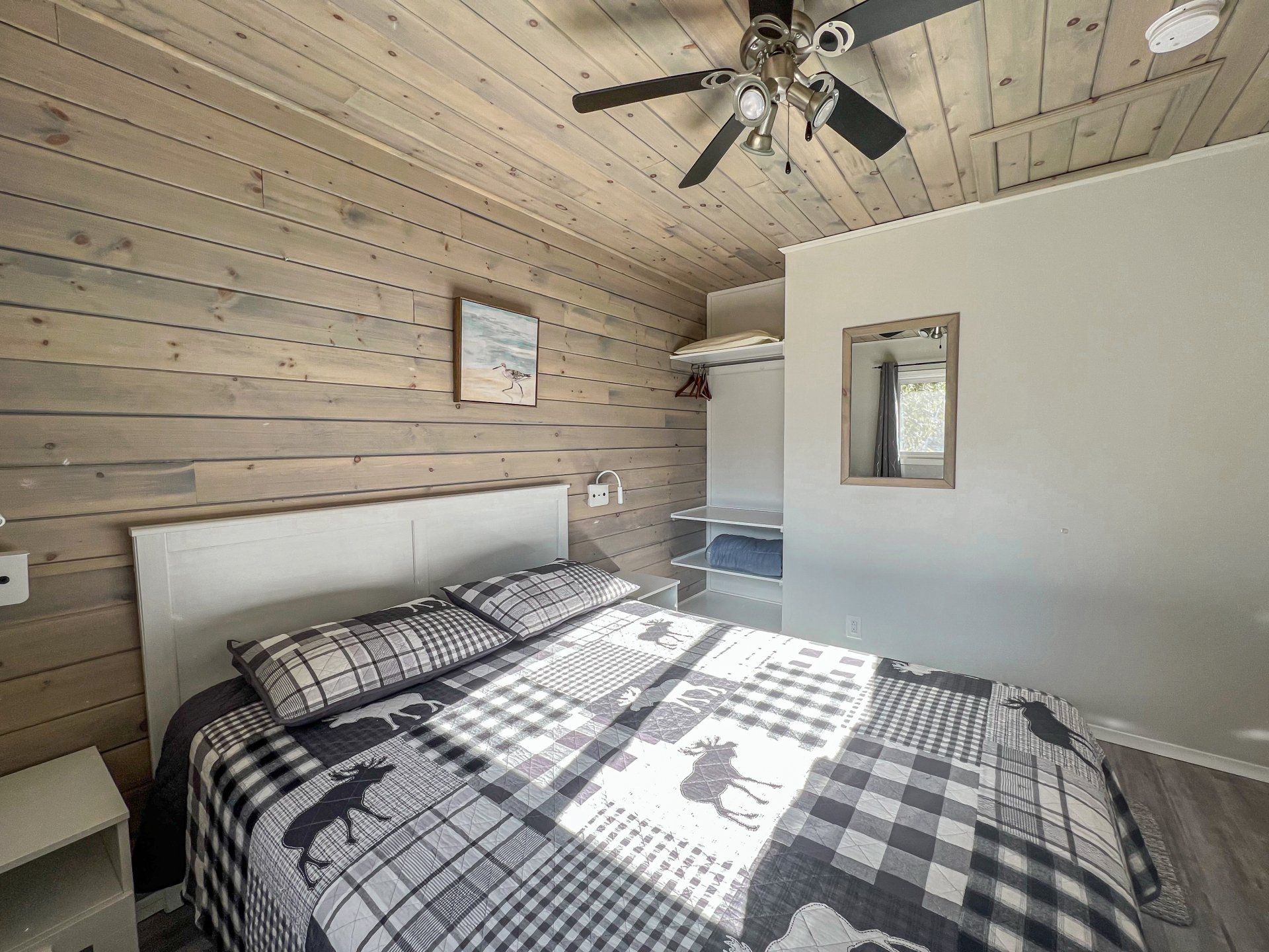 Cozy bedroom with wood-paneled walls, plaid bedspread, ceiling fan, and sunlight streaming through a window.