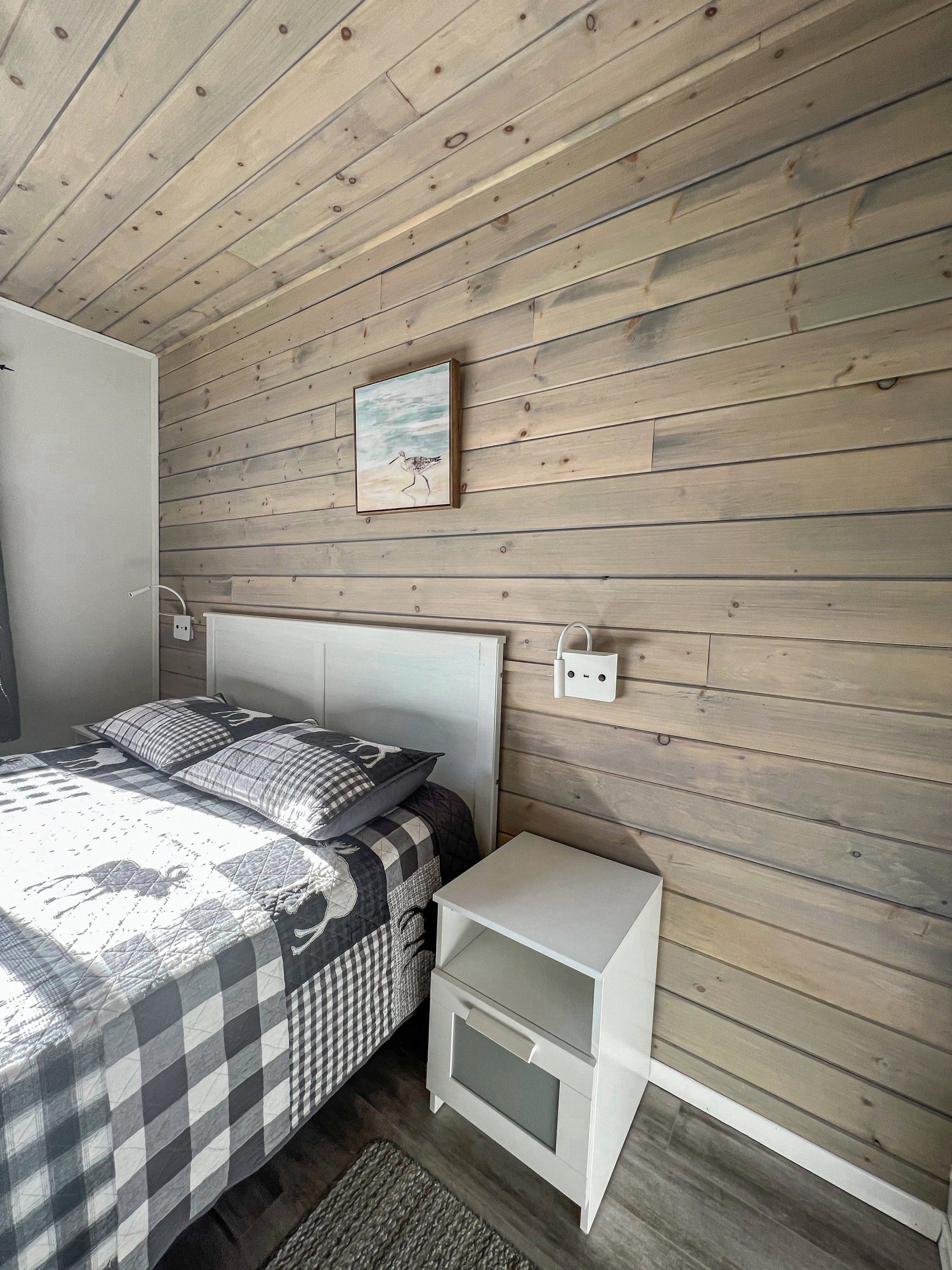 Small bedroom with gray-and-white plaid bed, wood-paneled wall, nightstand, and slanted ceiling