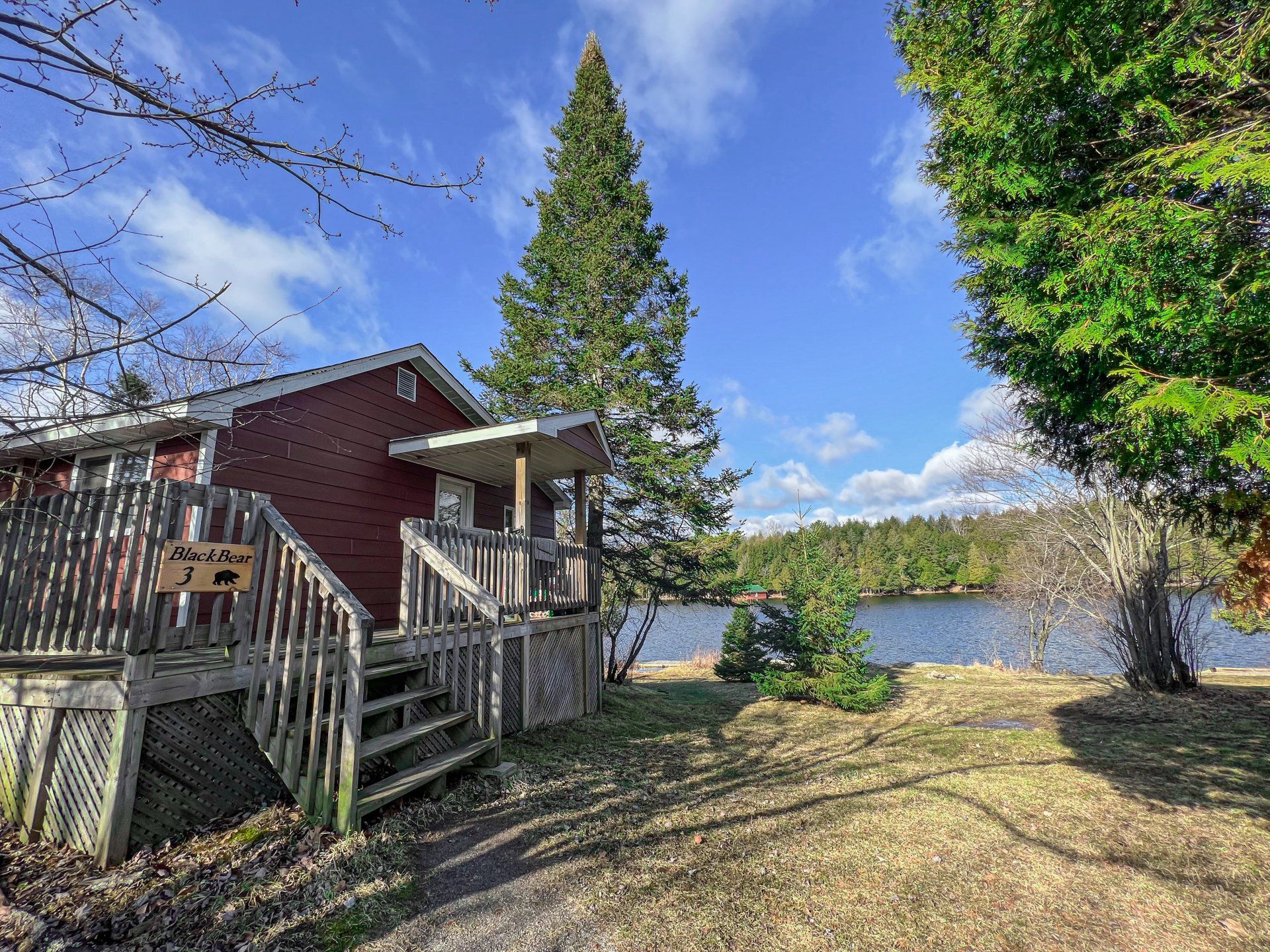 Red cabin with wooden deck by a lakeside under a blue sky, framed by trees and grass