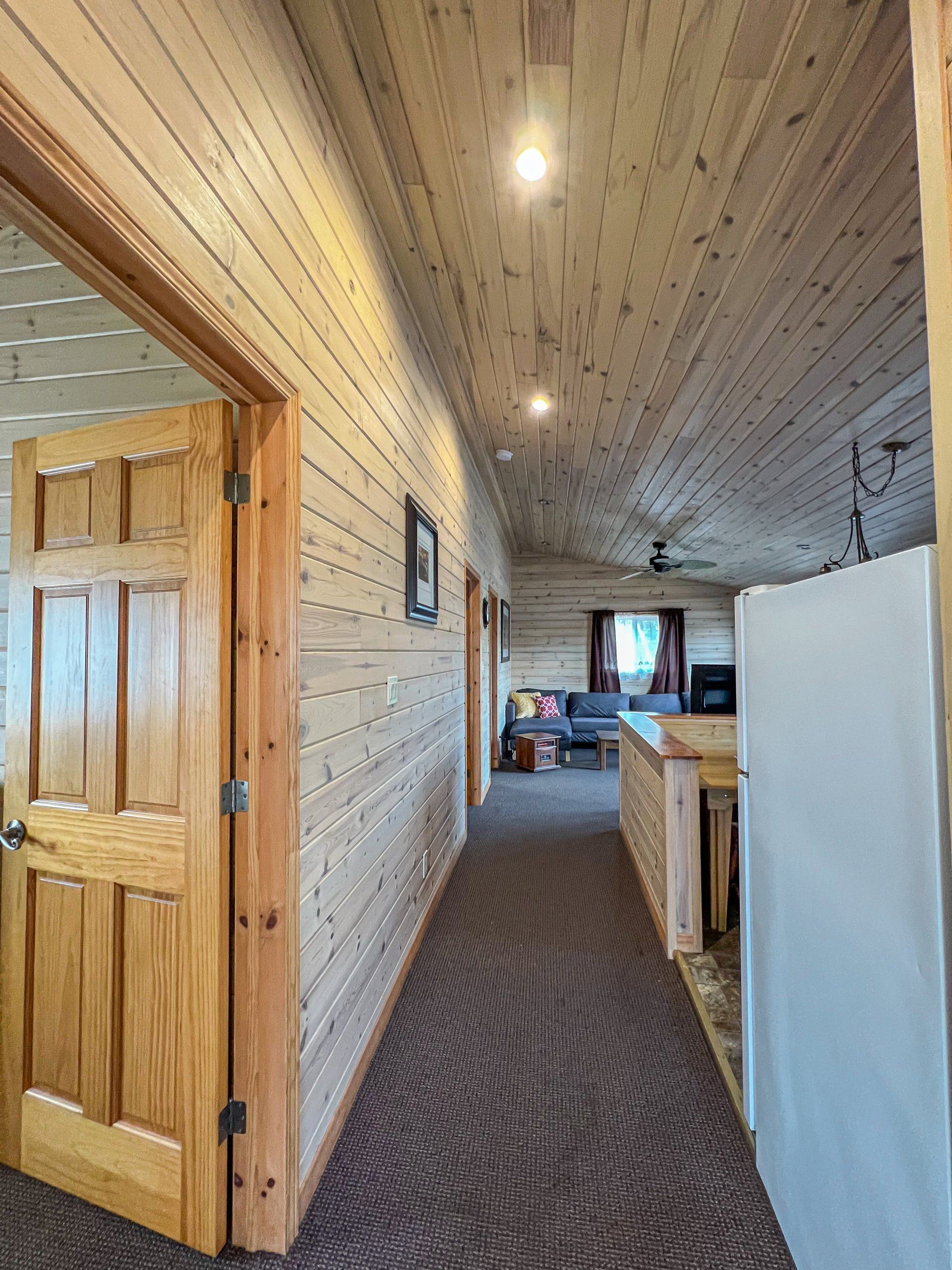 Narrow hallway with wood-paneled walls, carpet, open door, and kitchen area at the far end.