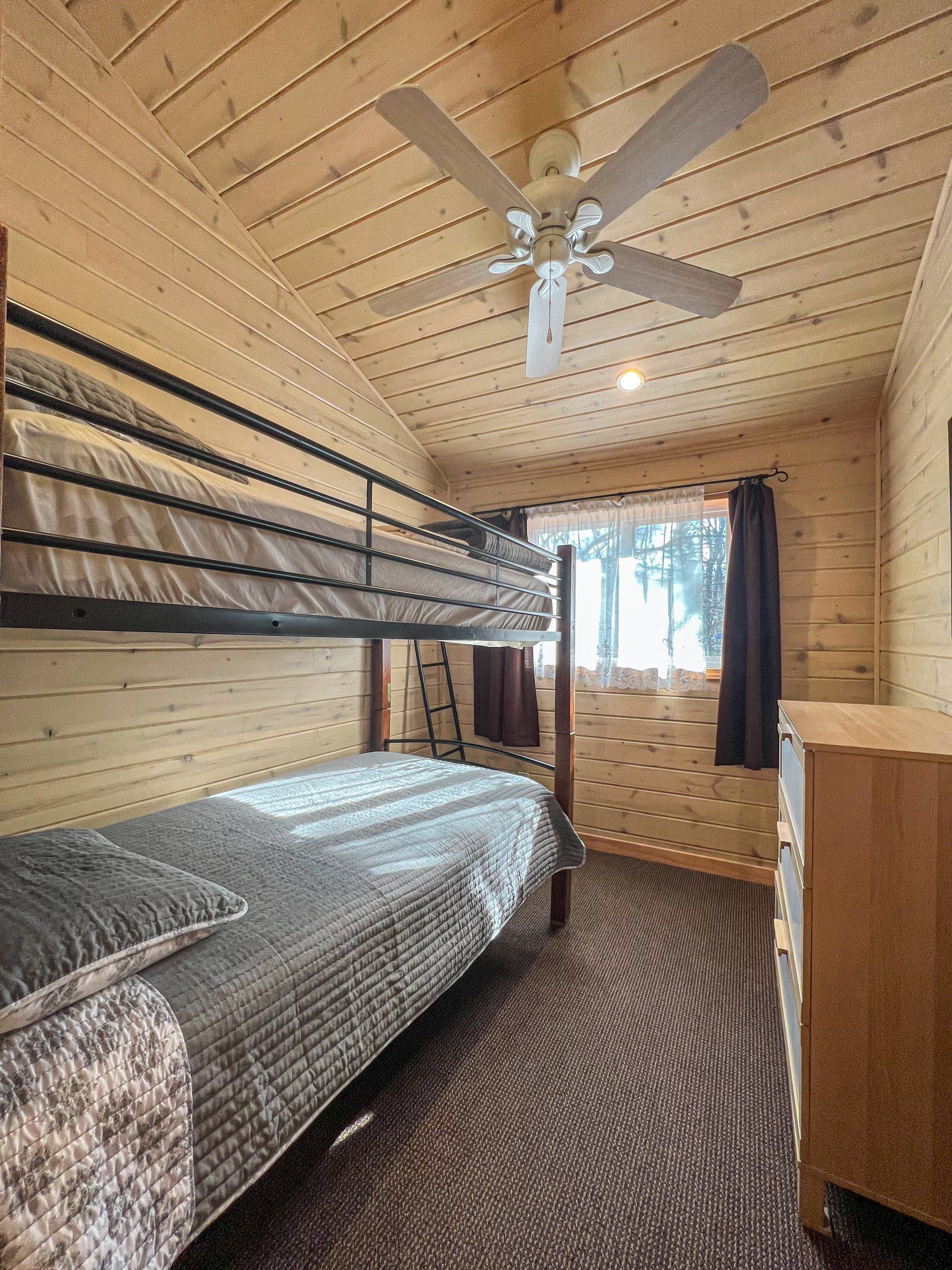 Small cabin bedroom with bunk beds, patterned carpet, ceiling fan, and window with curtains.