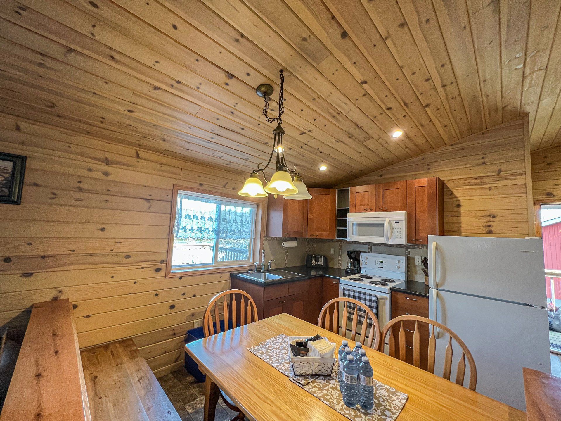 Wood-paneled kitchen and dining area with wooden table, hanging lights, white appliances, and window view