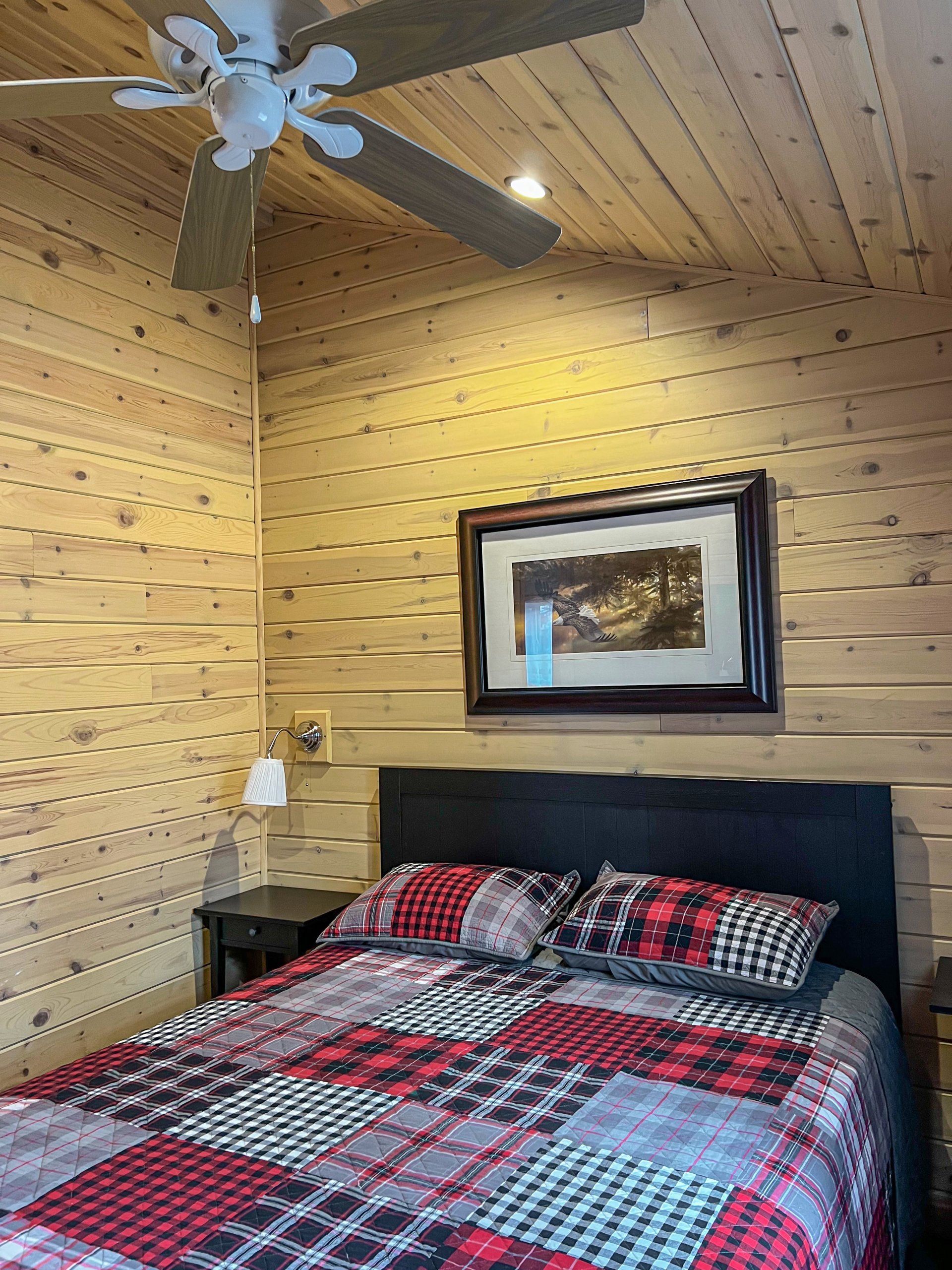 Cozy log cabin bedroom with plaid bedspread, black headboard, ceiling fan, and framed landscape art.