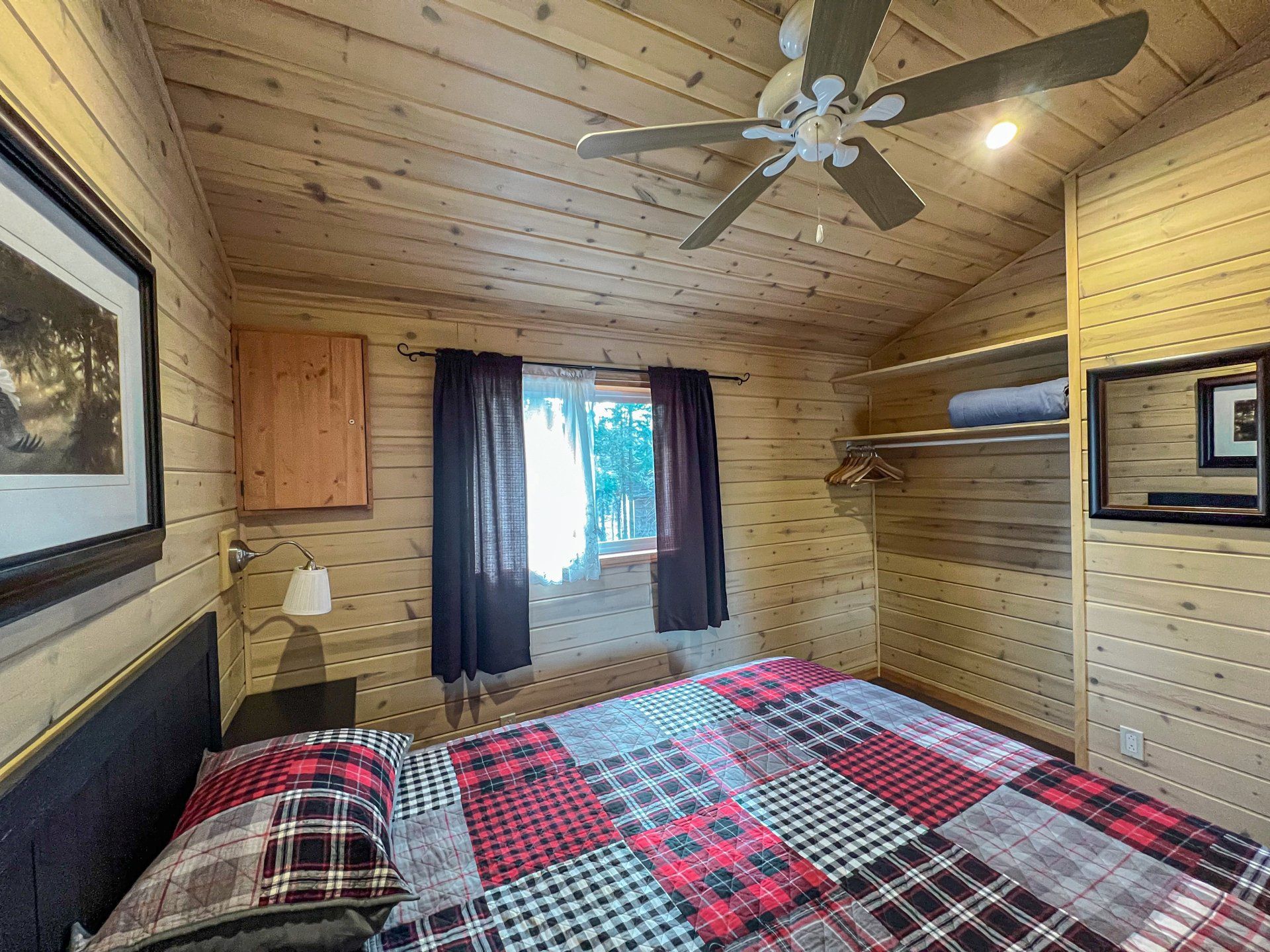Cozy wood-paneled bedroom with plaid bedspread, ceiling fan, window, and wall art