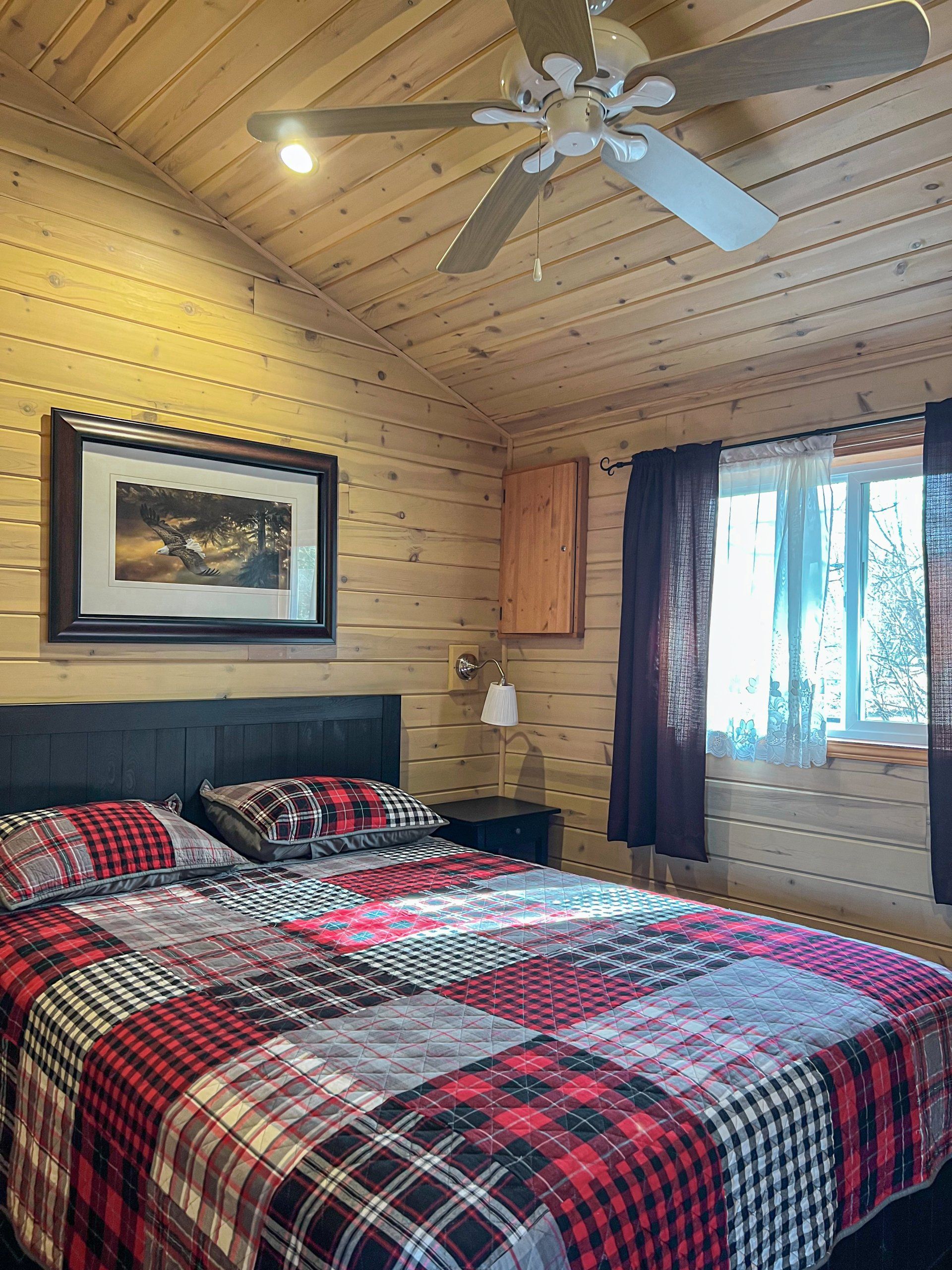 Cozy log cabin bedroom with plaid bedspread, wood-paneled walls, ceiling fan, and window light