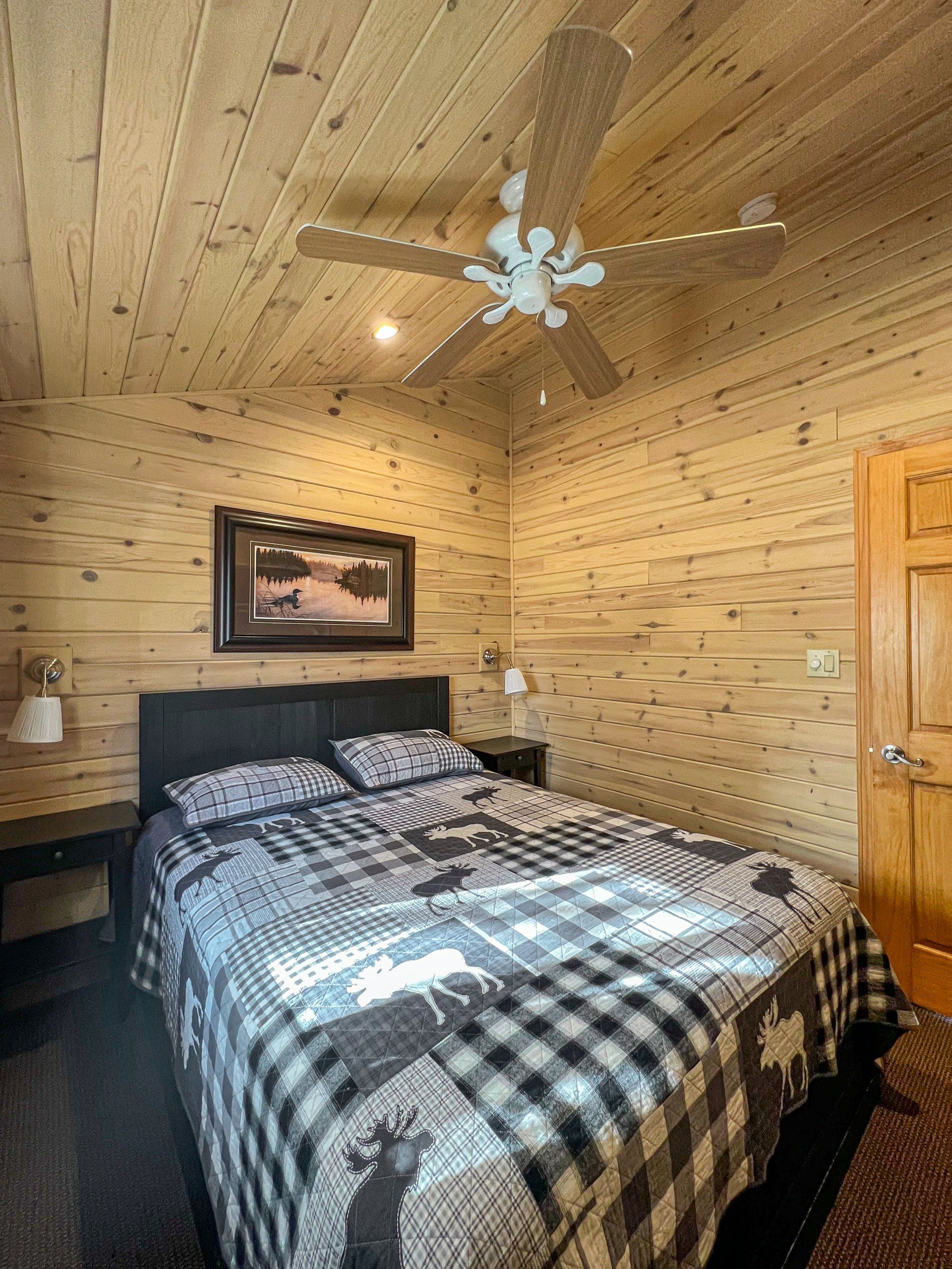 Rustic bedroom with wood-paneled walls, plaid bedspread, ceiling fan, and framed art above the bed