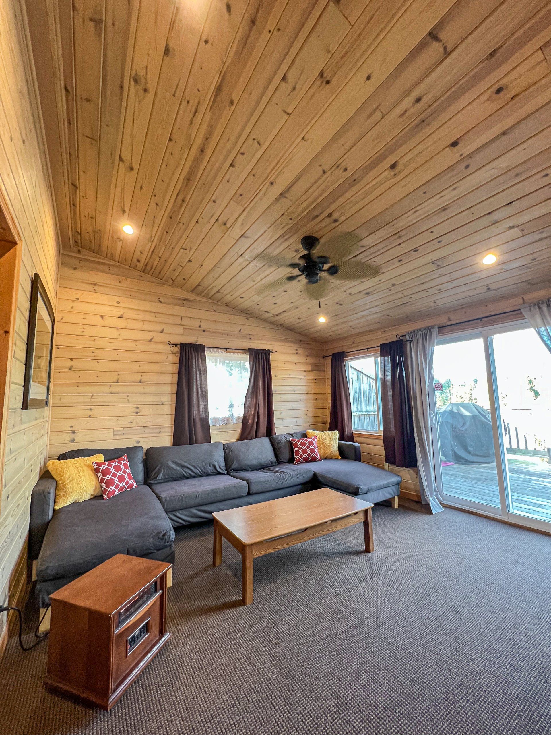 Cozy cabin-style living room with gray sectional sofa, wood ceiling, coffee table, and sliding glass doors